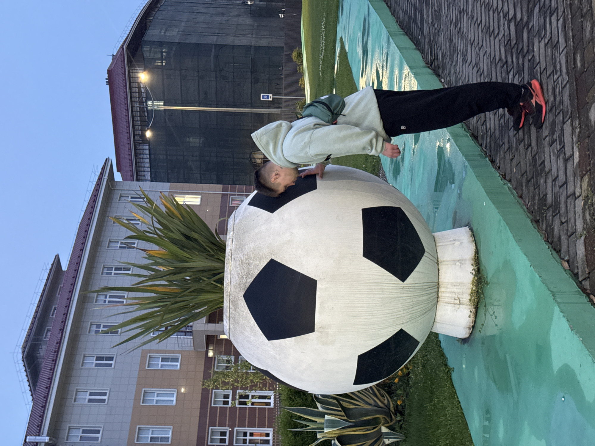 A person with a shaved head, wearing a light green hoodie, black pants, and red and black shoes, is resting their head and left hand on a large soccer ball-shaped art installation. The person has a green bag slung over their shoulder. The soccer ball is painted white with black pentagonal shapes, and a spiky green plant grows out of the top. The soccer ball rests on a short white pedestal and is surrounded by grass and other plants. The installation is on a turquoise-painted surface, which transitions to a brick path.
In the background, there is a multi-story building with a red roof and multiple windows. Further back, a larger, darker building is visible under a cloudy sky. The setting appears to be a park or public space in the city of Sirius, Russia.