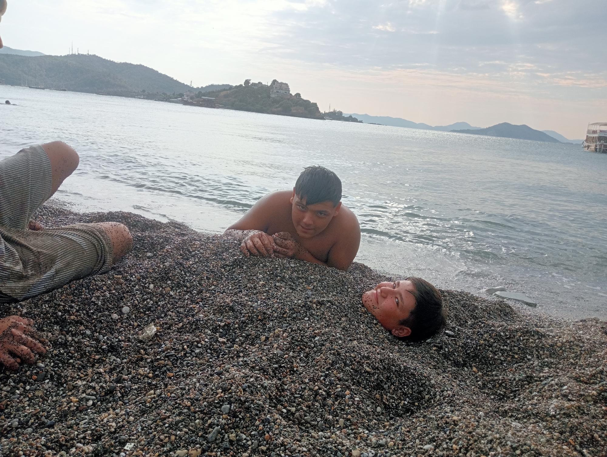 The image depicts two young males buried up to their necks in dark pebbles on a beach in Fethiye, Turkey. The sea is visible in the background, with a cloudy sky overhead. One of the boys is lying on his stomach with his hands resting on the pebbles in front of him, while the other is lying on his back with his head facing the camera. The water laps at the edge of the beach.