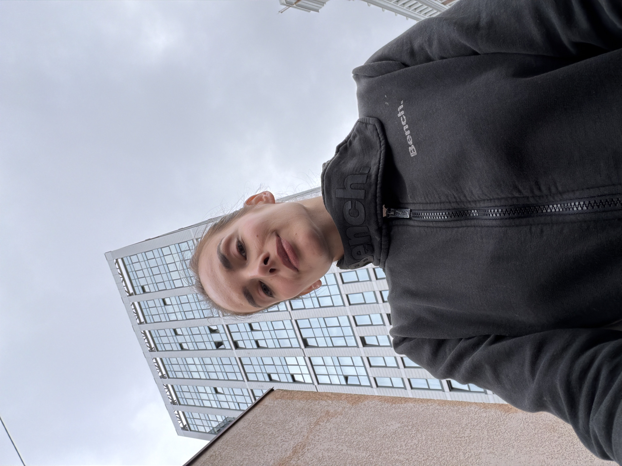 A low-angle, medium shot captures a woman from the chest up, taken outdoors in an urban environment. The woman, positioned slightly off-center to the right, is looking towards the camera with a neutral expression. She has light skin and her hair is pulled back. She is wearing a dark gray zip-up jacket with the word "Bench." visible in silver lettering on the right side of the chest, and also on the collar.
Behind the woman, the facade of a modern, multi-story building dominates the background. The building features a grid of numerous windows and a light-colored, textured exterior. To the left of this building is another building with a smooth, beige-colored facade, partially obscuring the taller structure. The sky above is overcast and appears gray. No specific landmarks definitively place the image in Ufa, Russia.