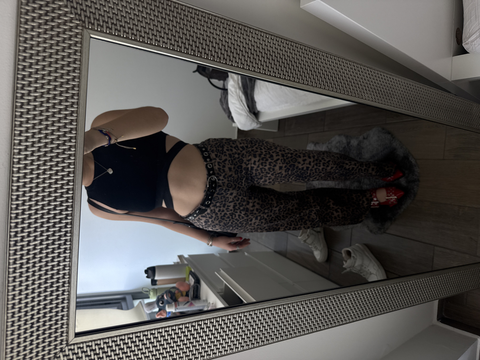 This media file captures a mirror selfie taken indoors, likely in a bedroom or dressing area, within Mexico City, Mexico. The primary image is rotated 90 degrees counter-clockwise.
The central subject is a person, visible from the torso downwards in the mirror's reflection. Their face is not visible. They are dressed in a black crop top featuring strap details, leopard print pants, and a black belt adorned with silver-toned eyelets and a round buckle. On their left wrist, multiple bracelets are visible, including a multi-colored string bracelet and a silver-toned chain. Red nail polish is present on their right hand. The person is wearing red high heels with buckle accents, standing on a fluffy grey faux fur rug.
The mirror is large and rectangular, encased in a distinctive frame with a textured, woven or braided pattern, appearing to be metallic in a silver-grey hue. The background reflected in the mirror reveals elements of the room: an unmade bed with white bedding and a grey blanket in the upper portion, and a wooden floor in the lower part. Also visible in the reflection are white shelving units or furniture, holding various objects such as bottles and containers. A pair of white sneakers rests on the wooden floor, outside the faux fur rug.
The image documents the act of taking a mirror selfie, likely to display the chosen outfit. There are no specific cues for the time of day, weather, or discernible emotions. No text is visible within the image.