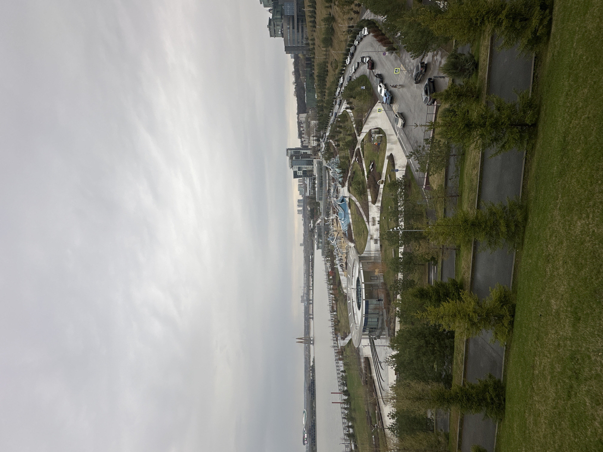 The image presents an elevated view of a cityscape under an overcast sky. A body of water, potentially a river or large canal, stretches through the scene, with buildings and infrastructure lining its banks. A park or public space is visible along the water's edge, featuring pathways, green areas, and possibly recreational facilities. Vehicles are observed on a road adjacent to the park.
This is likely Kazan, Russia, given the characteristic architecture and urban planning visible. The scene captures a blend of residential and commercial buildings, green spaces, and transportation routes, offering a snapshot of urban life in the city.