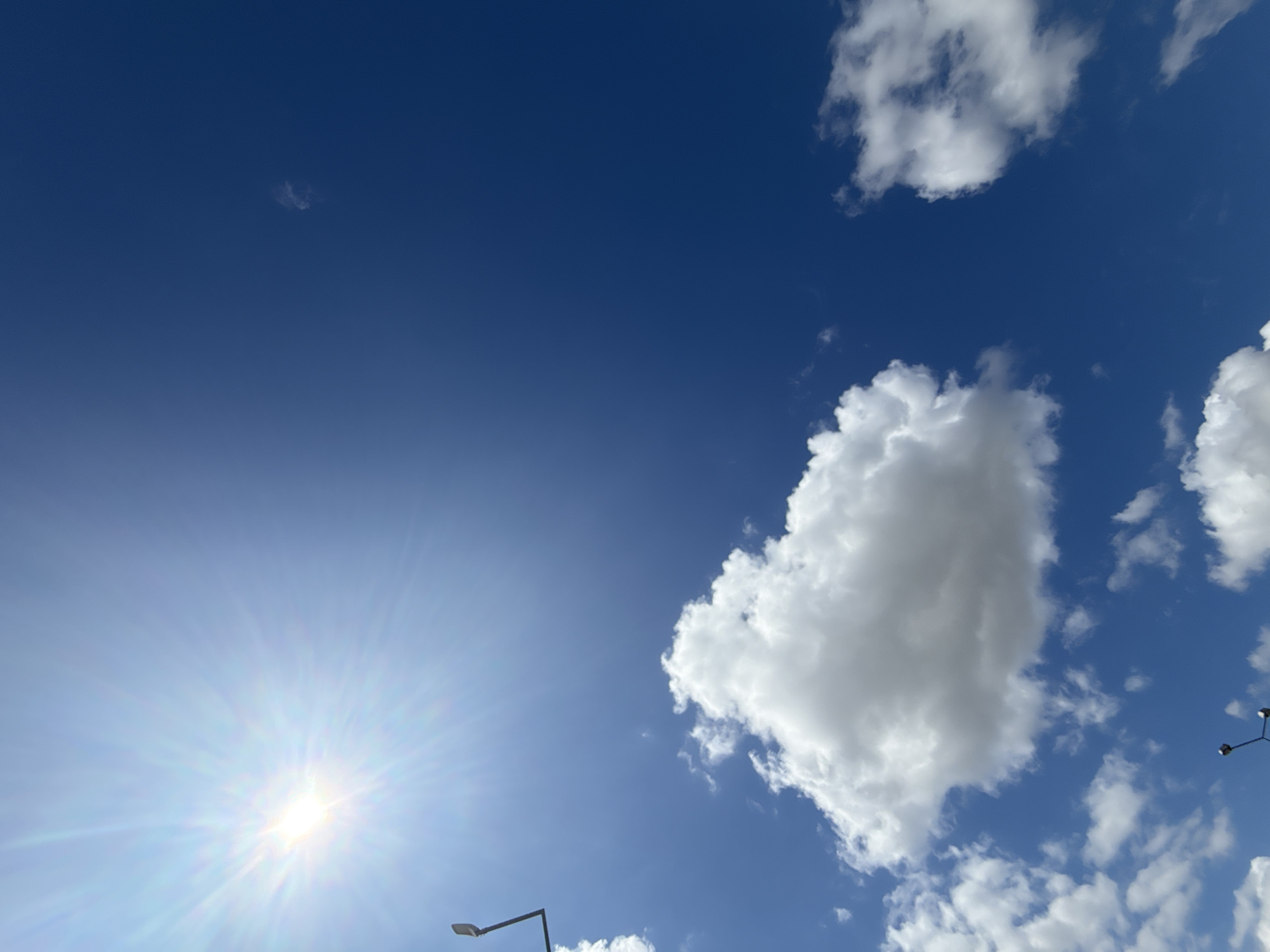 The image depicts a bright, sunny sky over the city of Żarska Wieś, Poland. The sun, a prominent source of light, is positioned in the upper left quadrant of the frame, casting a strong glare and visible rays. Large, fluffy white clouds are scattered across the deep blue sky. In the lower left portion of the image, a dark, angular structure, likely a lamppost or utility pole, is visible against the sky and clouds. There are no people or discernable activities taking place within the frame. The overall impression is that of a clear, fair-weather day.