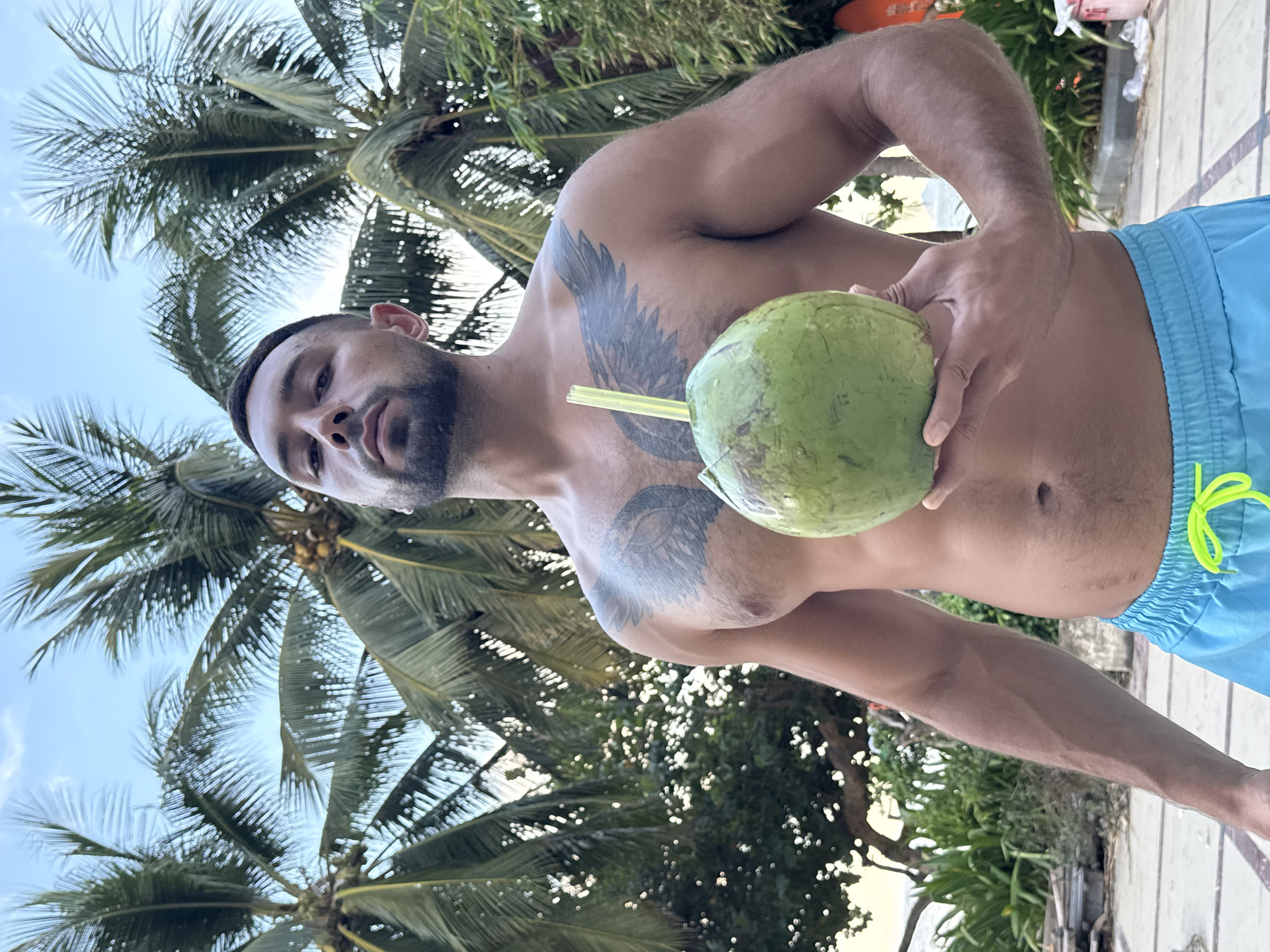 A man with a beard and tattoos on his chest is holding a green coconut with a straw in it. He is wearing blue swim trunks and is shirtless. The background consists of palm trees and a blue sky. The location is Nha Trang Ward, Vietnam.