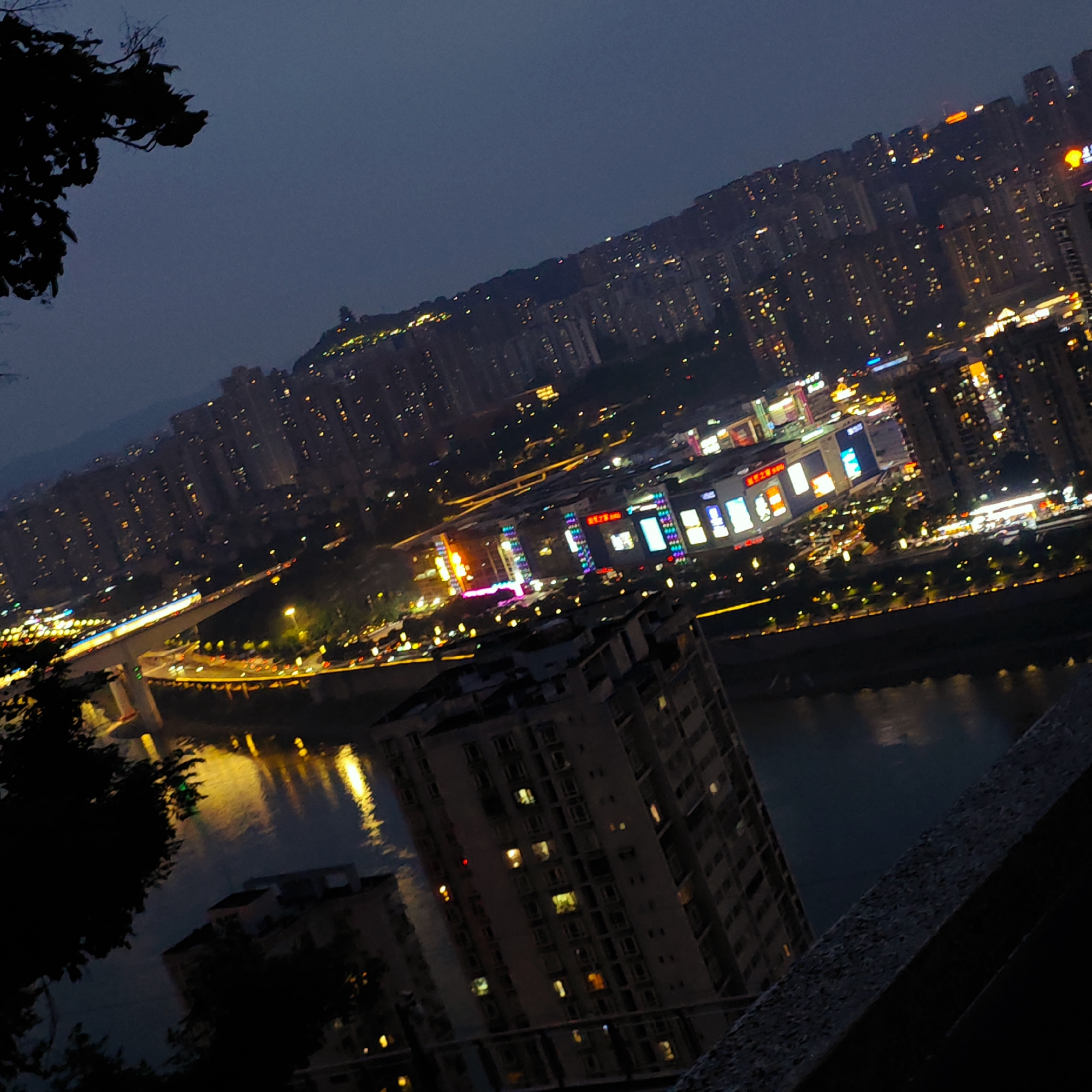 The image depicts a night view of the city of Yuzhong District, China, overlooking a river. The cityscape is densely packed with buildings illuminated by lights, reflecting on the water. A bridge spans the river, its lights adding to the urban glow. In the foreground, parts of buildings are visible, including an apartment complex. Vegetation is present in the lower left, framing the scene. A concrete structure is also visible in the foreground.