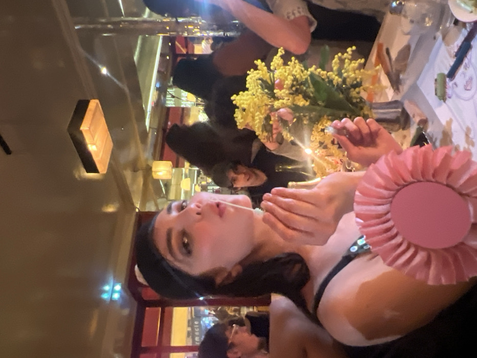 A woman in a black top and headband poses with a strand of spaghetti leading from her mouth. Several other people are present, seemingly dining or socializing. A bouquet of yellow flowers sits on the table beside tableware. Reflections indicate the setting is likely a restaurant, suggested by the lighting fixtures, red accents, and mirrored surfaces. A pink rosette is placed near the foreground.