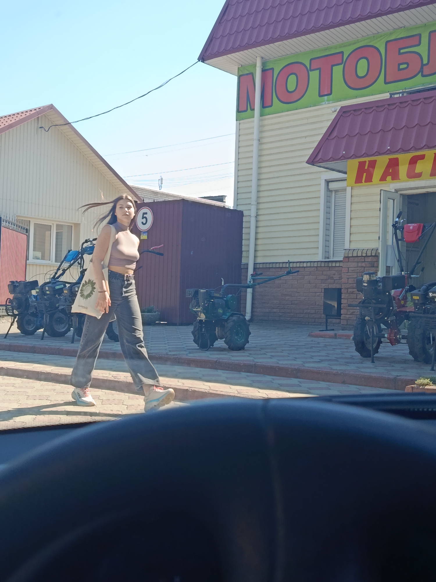 A young woman with long, dark hair is walking across a paved area in front of a building. She is wearing a sleeveless, light brown crop top, dark gray jeans, and light-colored sneakers. A light-colored tote bag with a sunflower design is slung over her shoulder. Behind her, several tillers are displayed outdoors, suggesting the location is a store selling agricultural equipment. The building behind the tillers has a yellow-paneled exterior with red trim and a red tiled roof. A sign with large red letters on a green background is visible on the building, reading "МОТОБ..." and another sign with red letters on a yellow background reads "HAC". To the left of the woman, a white building with a similar red-tiled roof is visible. A circular sign with the number "5" on it is mounted on a dark brown post behind the woman. The scene is brightly lit by sunlight, indicating it is daytime, and the sky is clear and blue. The woman appears to be walking with a purposeful stride, and her hair is being blown by a gentle breeze. The overall impression is of a sunny day in a commercial area.