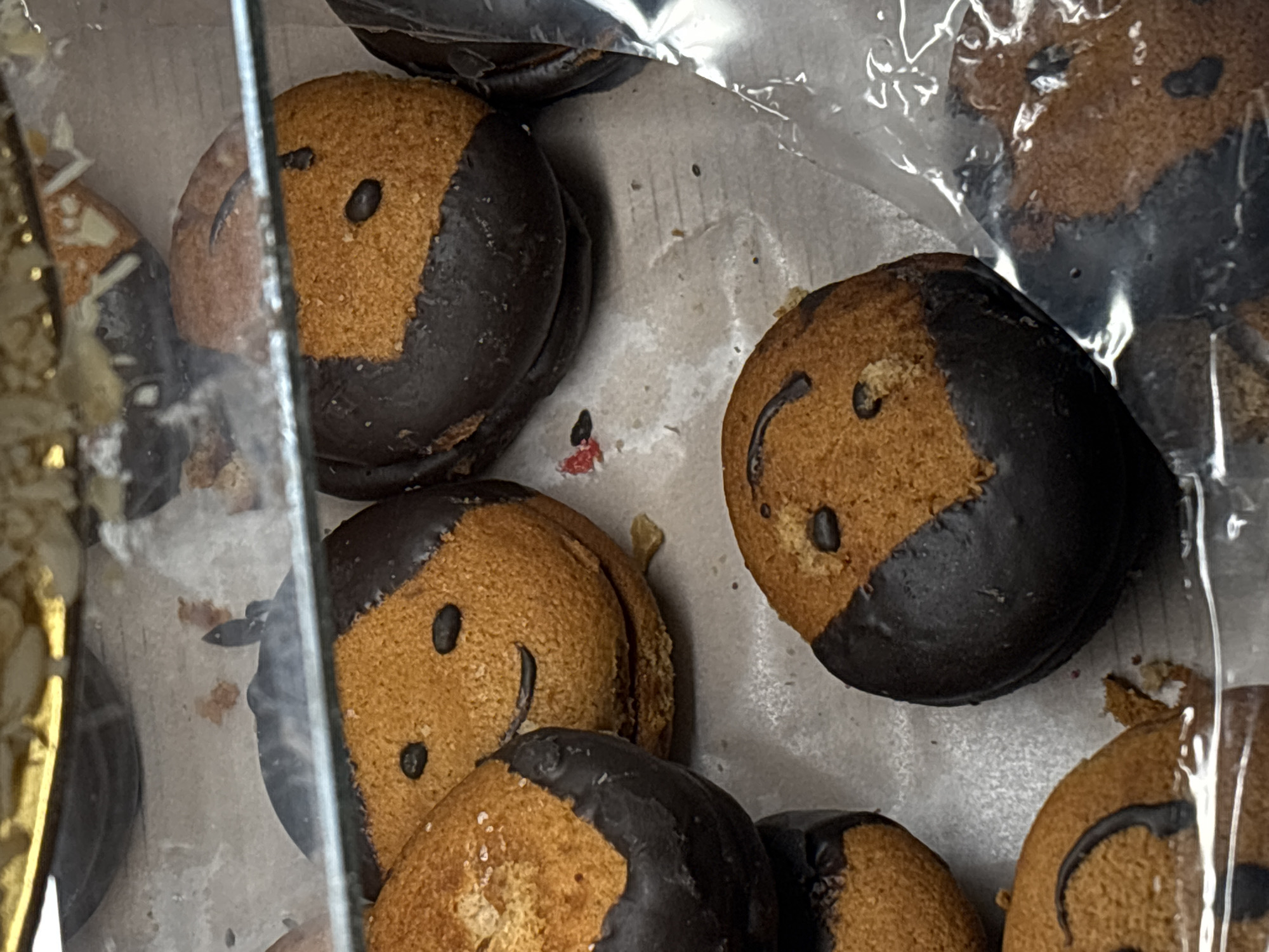 The image shows a selection of cookies inside a display case, likely in a bakery or confectionary shop. The cookies are round and have a two-toned appearance, with one half a golden-brown color and the other half covered in dark chocolate. Each cookie has a simple "face" design on the golden-brown part, consisting of a small oval for the nose and a curved line for the mouth. The cookies are arranged in a box, possibly lined with paper. The location is unknown.