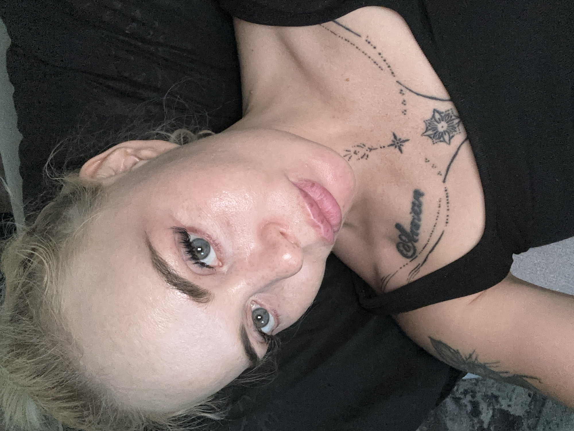 A close-up, high-angle shot focuses on the face and upper chest of a woman with blonde hair pulled back. She has light blue eyes, noticeable eyelashes, and fair skin with visible pores and some shine. Her lips are slightly parted, and she has a tattoo on her chest. The background is dark and indistinct, suggesting a dimly lit indoor environment.