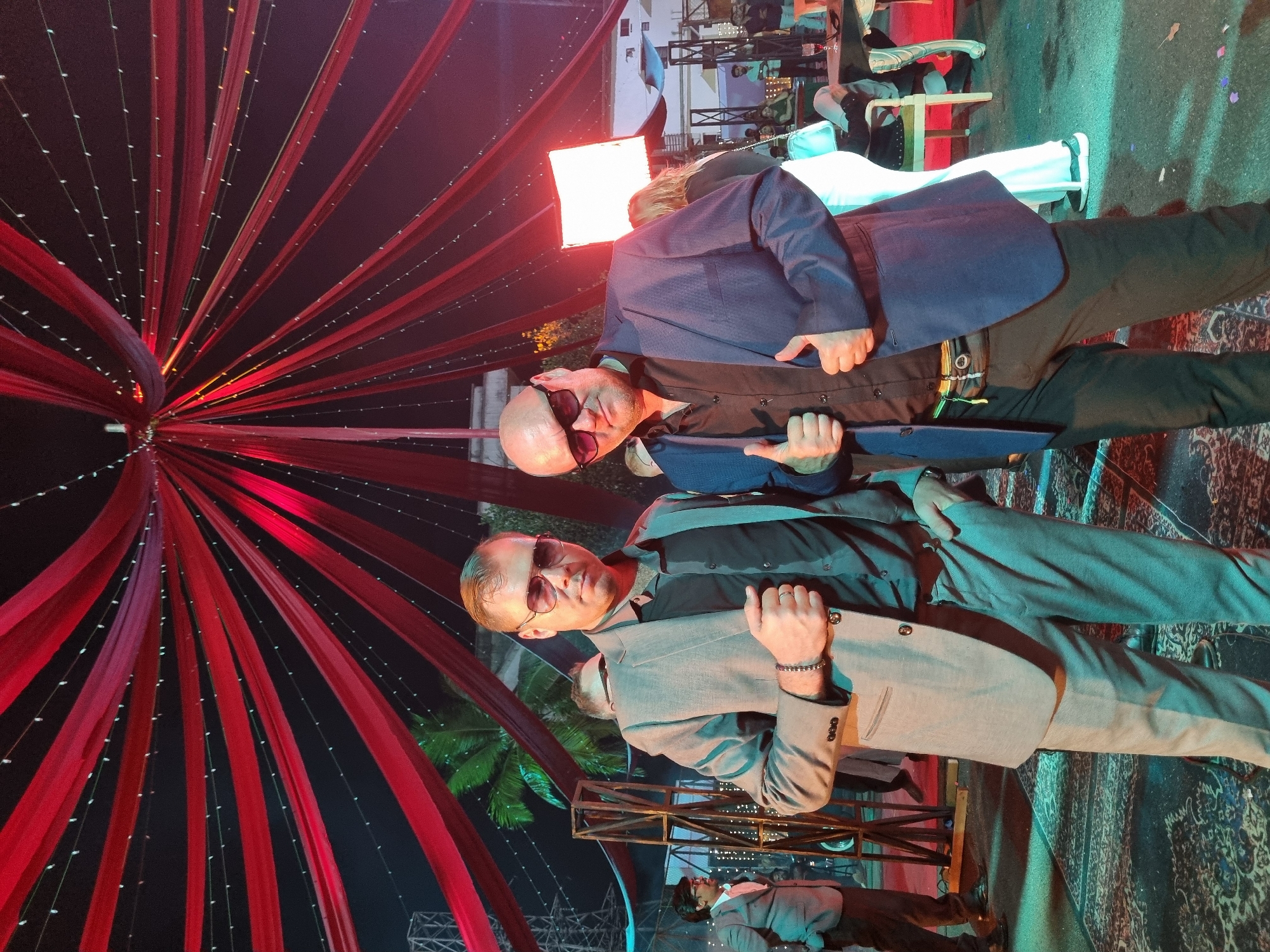 The image captures two men posing at what appears to be an evening event or celebration. In the foreground, two men stand side-by-side, facing the camera. The man on the left (when viewed upright) is bald, wearing sunglasses, a dark shirt, and a grey blazer, with a metal bracelet on his right wrist. The man on the right (when viewed upright), also bald and wearing sunglasses, sports a dark shirt under a blue blazer and dark trousers. Both have their hands positioned casually, creating a relaxed yet confident demeanor.
The setting is characterized by elaborate decorations, featuring striking red fabric drapes suspended overhead in a radial pattern, resembling a tent or canopy. These drapes are interwoven with strings of small, white fairy lights that emit a soft glow against the dark background. The scene is illuminated by these decorative lights, as well as ambient blue and green stage lighting in the background, indicating it is nighttime. A bright, rectangular white light panel is visible behind the man in the blue blazer, suggesting professional lighting for the event.
The ground is covered with a patterned rug. In the background, other event attendees, chairs, tables, and structural elements like metal scaffolding are faintly visible. The presence of potted plants, possibly palm trees, suggests an outdoor or semi-outdoor venue. The overall atmosphere appears festive and social. There is no discernible text visible in the image.