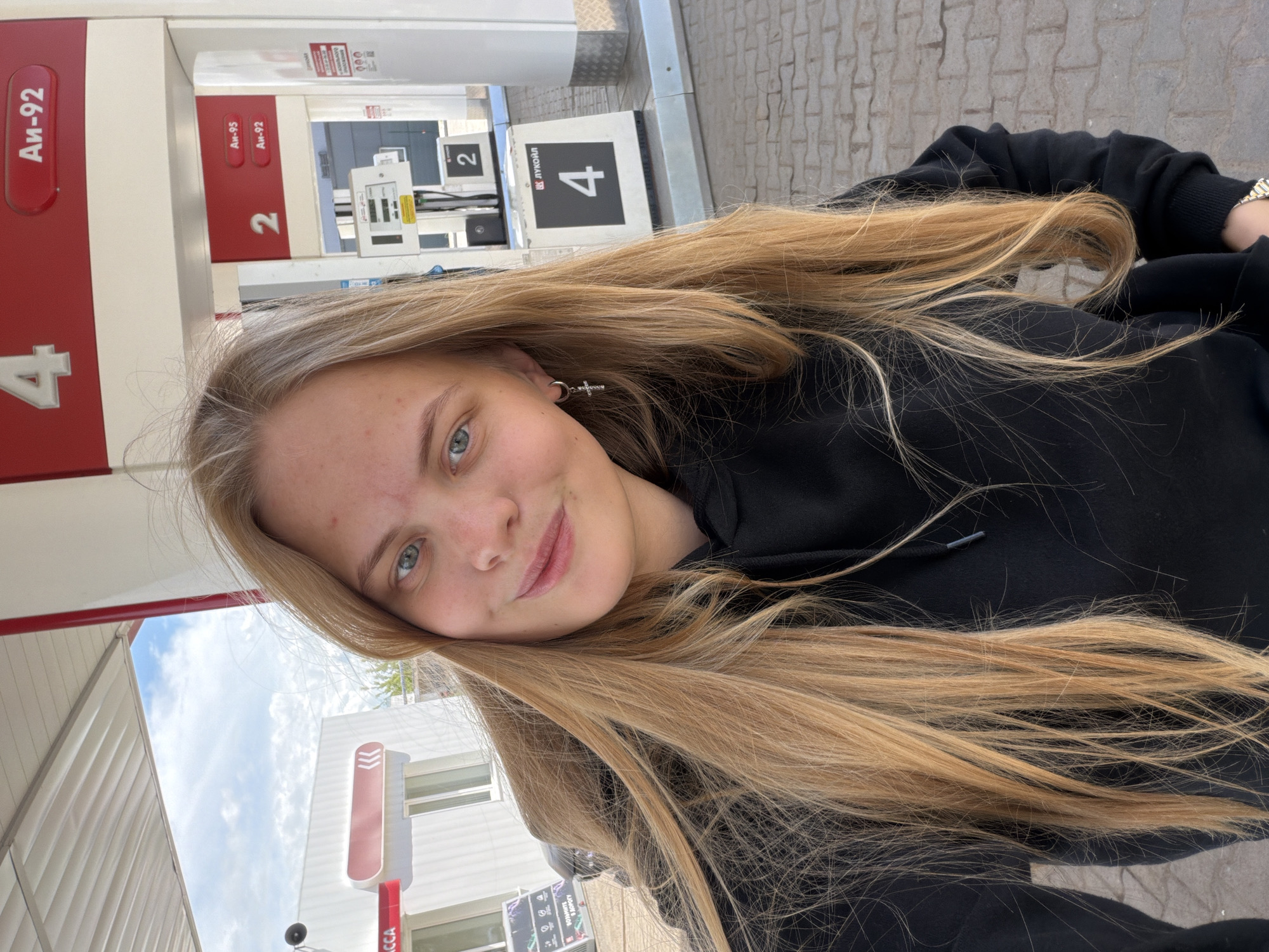 The image shows a woman with long, blonde hair wearing a black hoodie. She is standing at a Lukoil gas station, as indicated by the signage visible in the background. The gas station features pumps with numbers "2" and "4", as well as signs indicating the availability of "Аи-92" and "Аи-95" gasoline. The woman appears to be taking a selfie. The image was taken in the city of Chaikovsky, Russia.