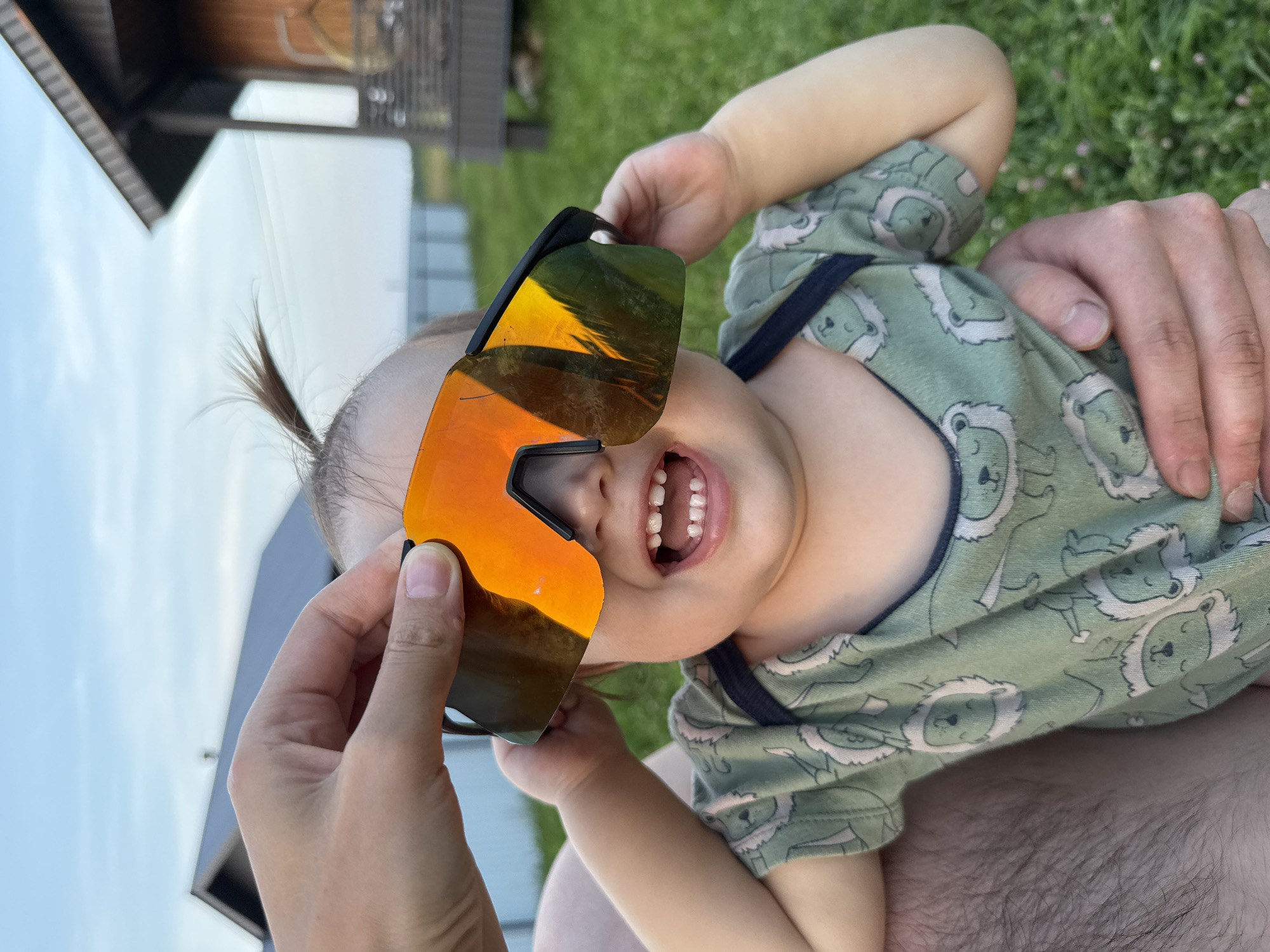 A close-up, vertical outdoor shot shows a young child wearing a green onesie with a repeating lion pattern. The child is being held by an unseen adult and is laughing with their mouth open, revealing small teeth. A person's hand holds a large pair of orange-tinted wraparound sunglasses over the child's eyes, obscuring their vision and reflecting a sunset scene. The background is blurred but appears to be a grassy field with a wooden structure and a fence. The image is taken in the village of Podshivalovo, Russia.