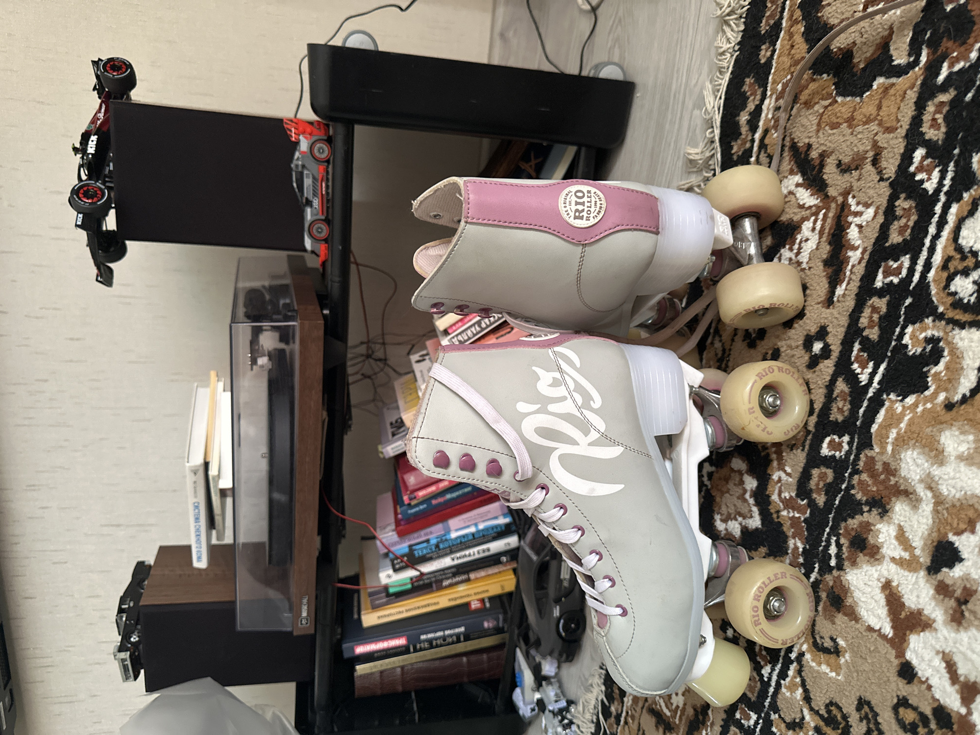 The image displays a pair of light gray and pink roller skates positioned on an oriental rug in a domestic setting. The roller skates have white wheels with pink accents and "Rio Roller" branding. Behind the roller skates, a bookshelf filled with books is visible, and on top of the shelf, there are several model cars and what appears to be a record player. The background shows a wall with a textured finish and some electronic equipment, including speakers. The scene is indoors, and the lighting suggests it is daytime. There are no people or animals present in the image. There is no information in the image to suggest it is located in Krasnodar, Russia.