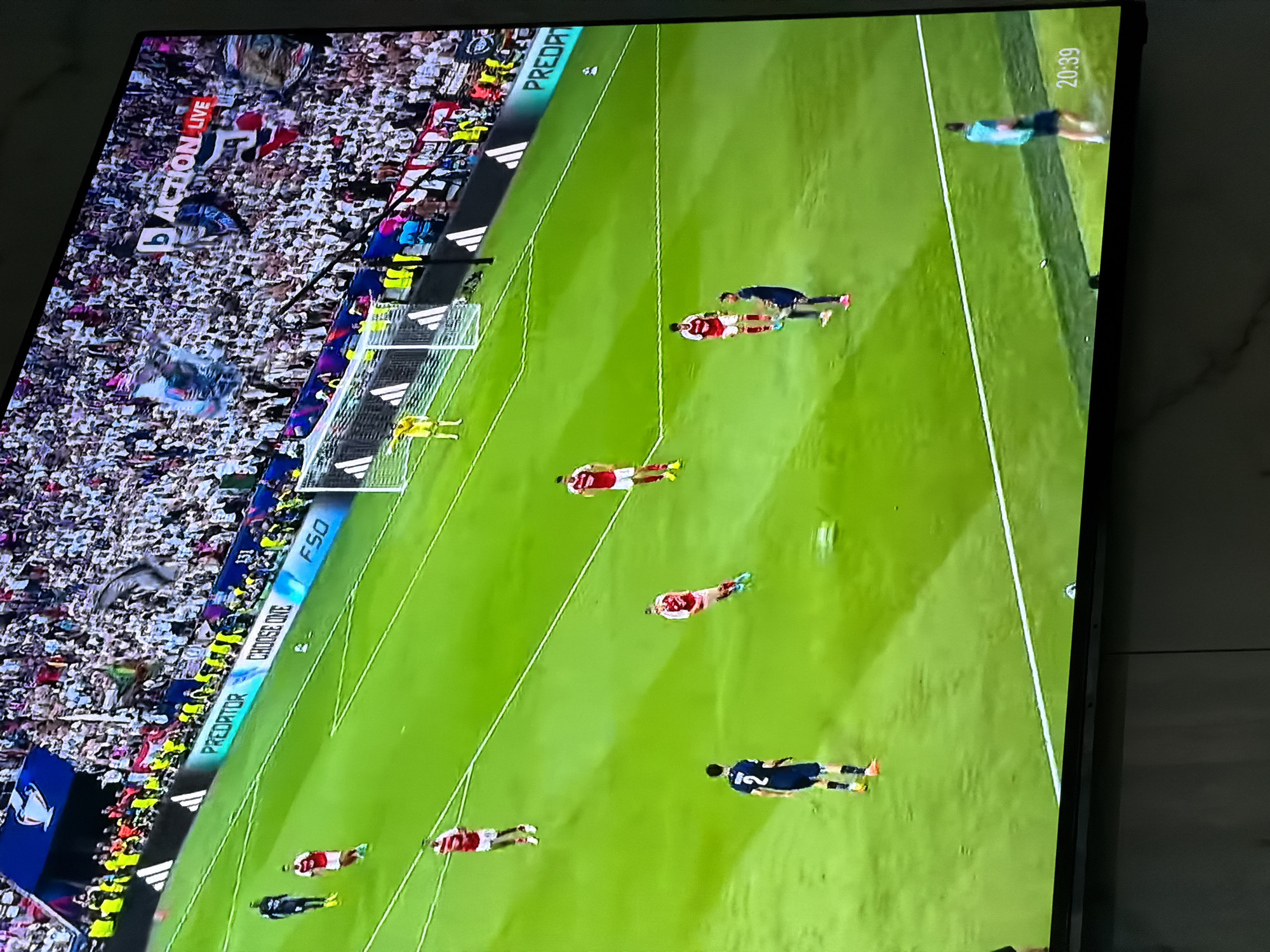 The image shows a televised soccer game, likely viewed on a flat-screen television. The perspective is from an elevated angle, looking down at the screen.
The televised soccer match appears to be a high-profile game with a large crowd of spectators. The game features two teams; one in red and white jerseys, and the other in navy blue. The players are positioned across the green soccer field. The time displayed on the upper right corner of the television screen reads "20:39."
The image provides no information to support the assertion that the location is in Asenovgrad, Bulgaria.