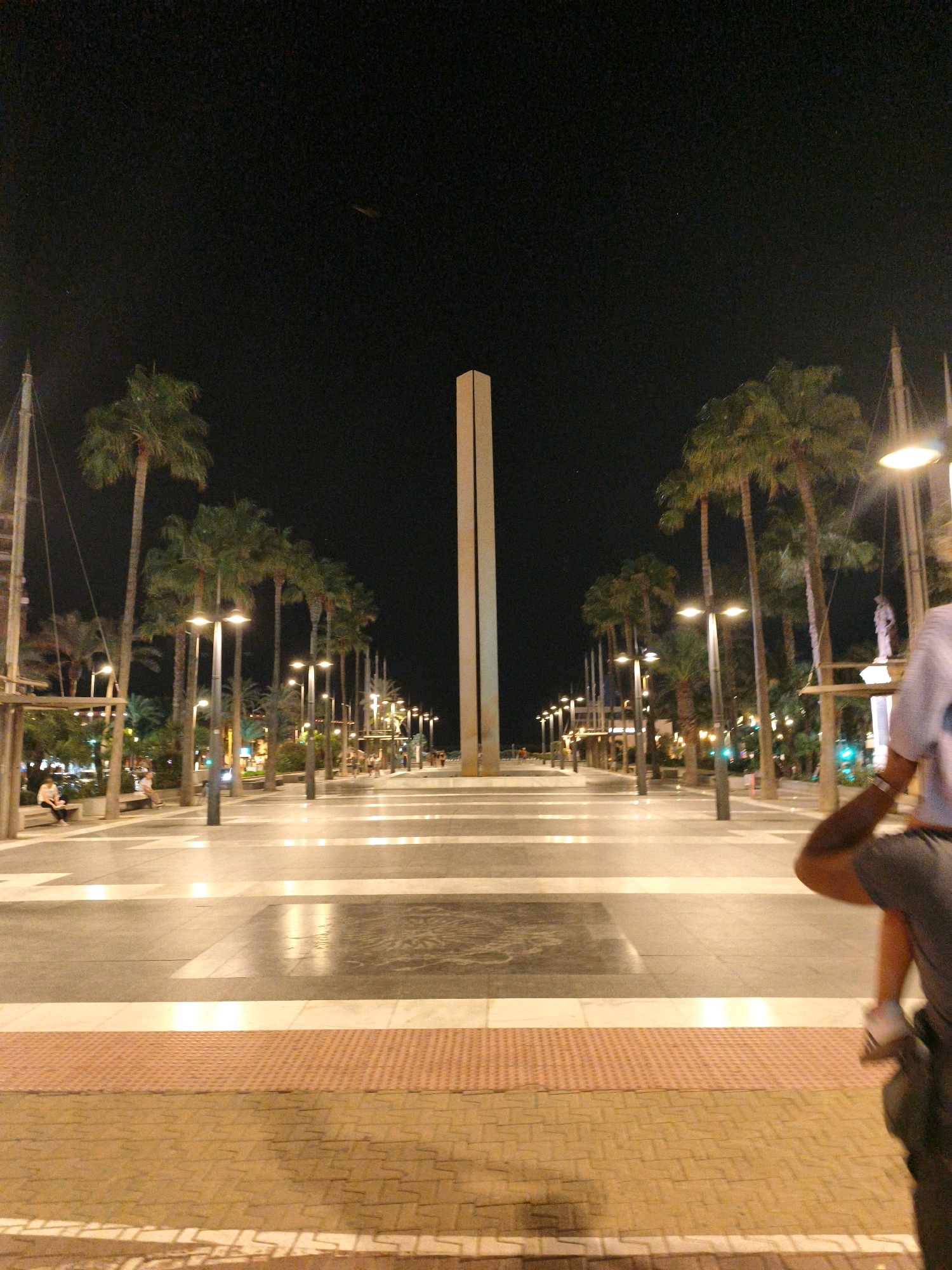 The image depicts a nighttime scene on a wide, paved promenade in Almería, Spain, likely near the Paseo Marítimo. The promenade is lined with tall palm trees on the left and right sides, illuminated by streetlights. In the center of the promenade, a tall, slender obelisk-like structure stands prominently against the dark night sky. The ground is paved with large rectangular tiles, with reflective strips running horizontally across the promenade.
In the foreground, a person, only partially visible from the back and right side, is in motion, suggesting they are walking or running. They are wearing a dark top and dark shorts, with one leg extended. The rest of the promenade shows several individuals seated on benches near the palm trees on the left, observing the scene or resting. Further down the promenade, more streetlights and structures are visible, indicating the presence of shops or other establishments. The overall atmosphere is that of a lively urban promenade at night.