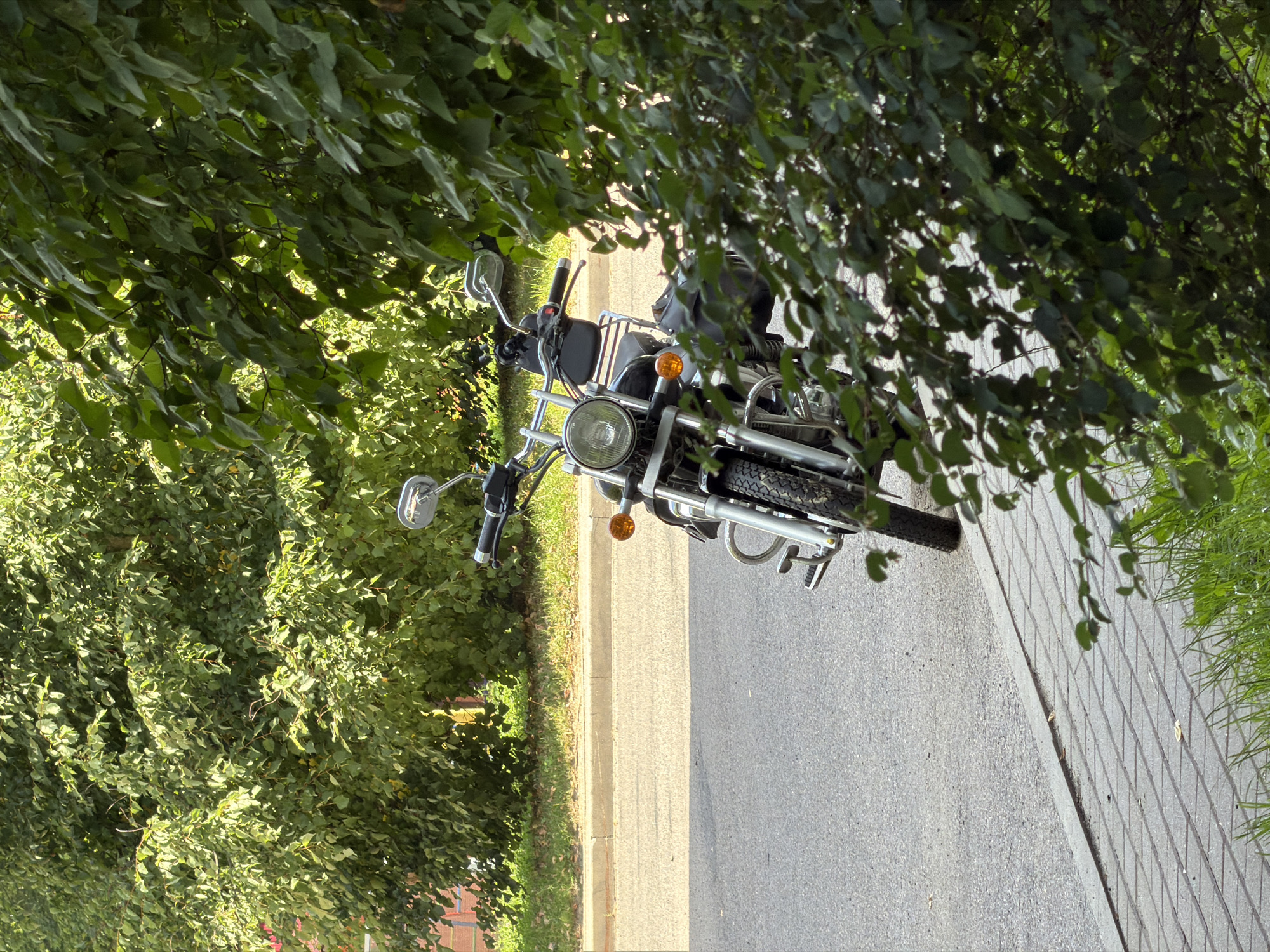A chrome and black motorcycle is parked on the side of a paved street, its front tire and handlebars visible. The motorcycle is positioned at an angle to the curb, with the front wheel angled towards the road. A sidewalk made of grey interlocking bricks runs alongside the street. Lush green trees with dense foliage overhang the motorcycle and the sidewalk, casting shadows and partially obscuring the view of a building in the background. The scene appears to be in a residential area within Minsk, Belarus, given the common elements of urban landscaping and street infrastructure found in the city. The lighting suggests it is daytime, with sunlight filtering through the leaves.