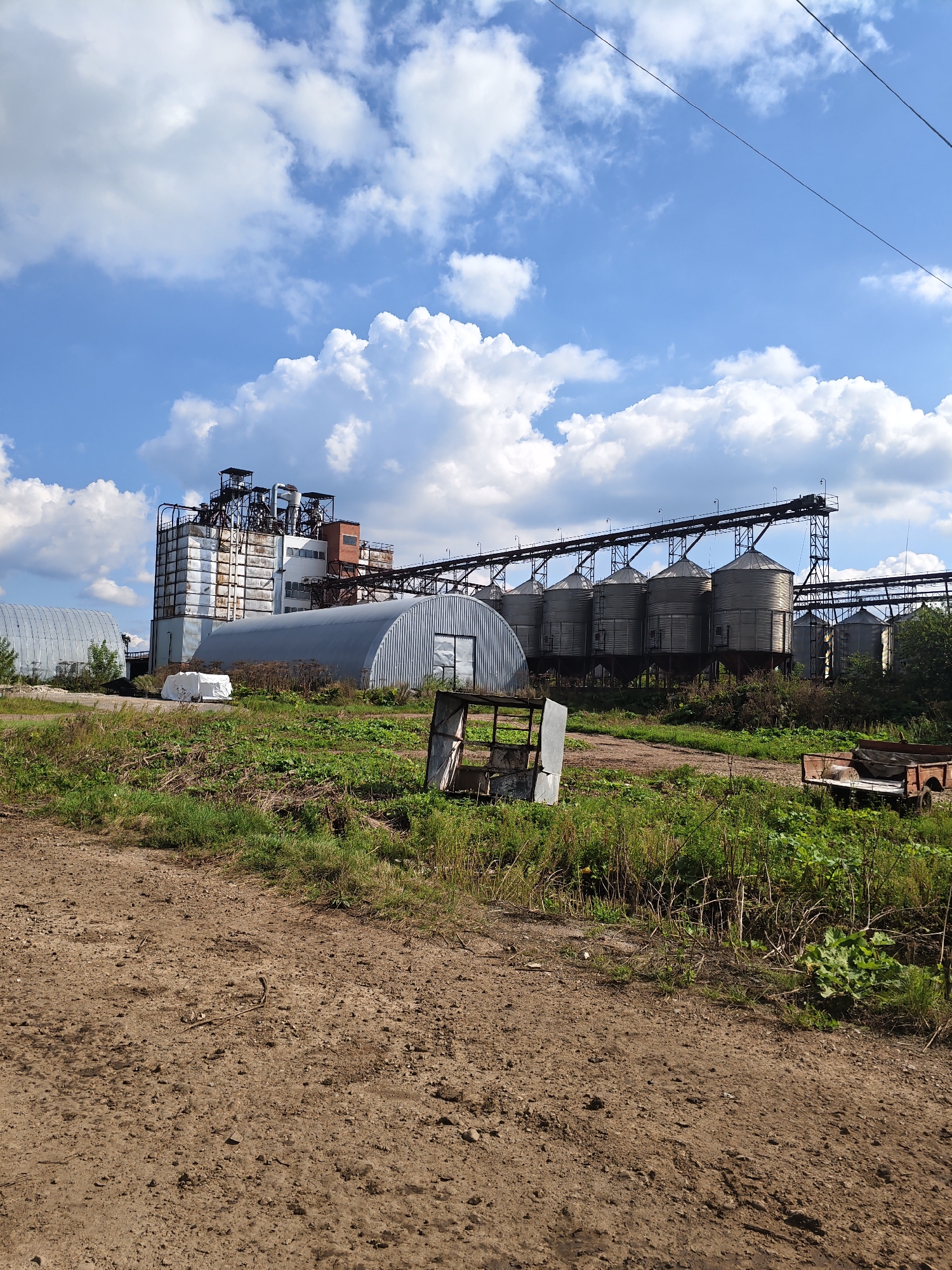 The image depicts an industrial site with a clear blue sky and scattered white clouds. In the background, a multi-story industrial building with a metallic exterior is visible, along with a series of tall, cylindrical storage silos connected by an overhead conveyor system. To the left of the main building, a large, curved, silver-colored structure, possibly a hangar or another storage facility, is present. In the foreground, a dirt path leads through a grassy area with scattered debris, including what appears to be a small, dilapidated metal shelter and a rustic trailer. The environment suggests a rural or semi-industrial setting.