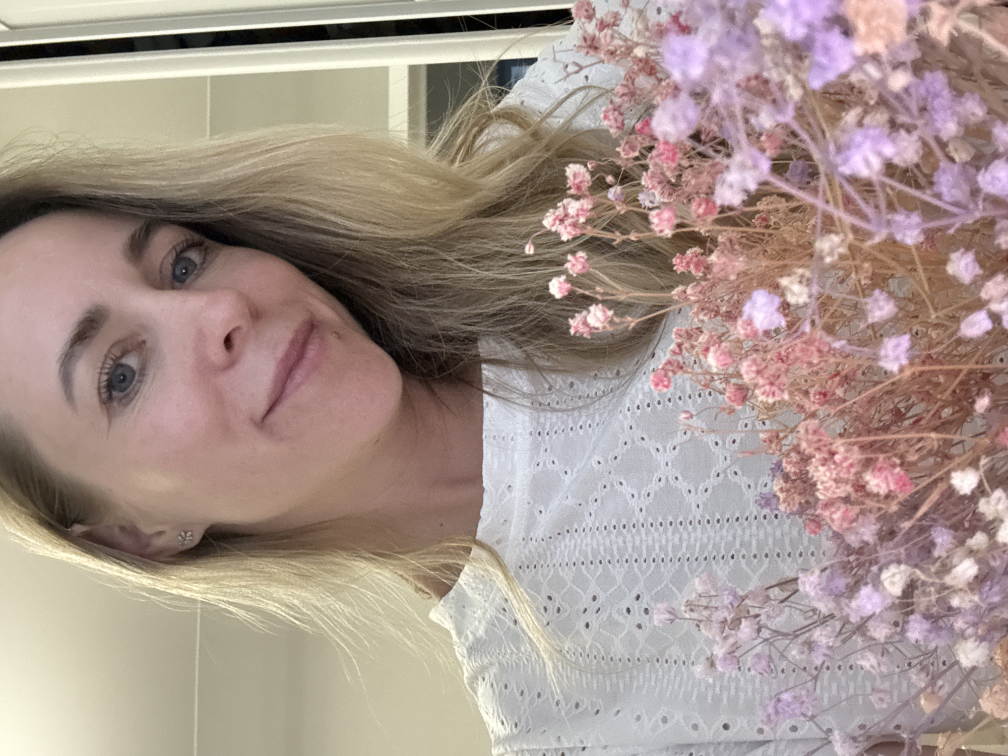 A woman with blonde hair and blue eyes is holding a bouquet of pink and purple flowers. She is wearing a white eyelet top. The background appears to be an interior with light-colored walls. There is no information to suggest the location is Chaikovsky, Russia.