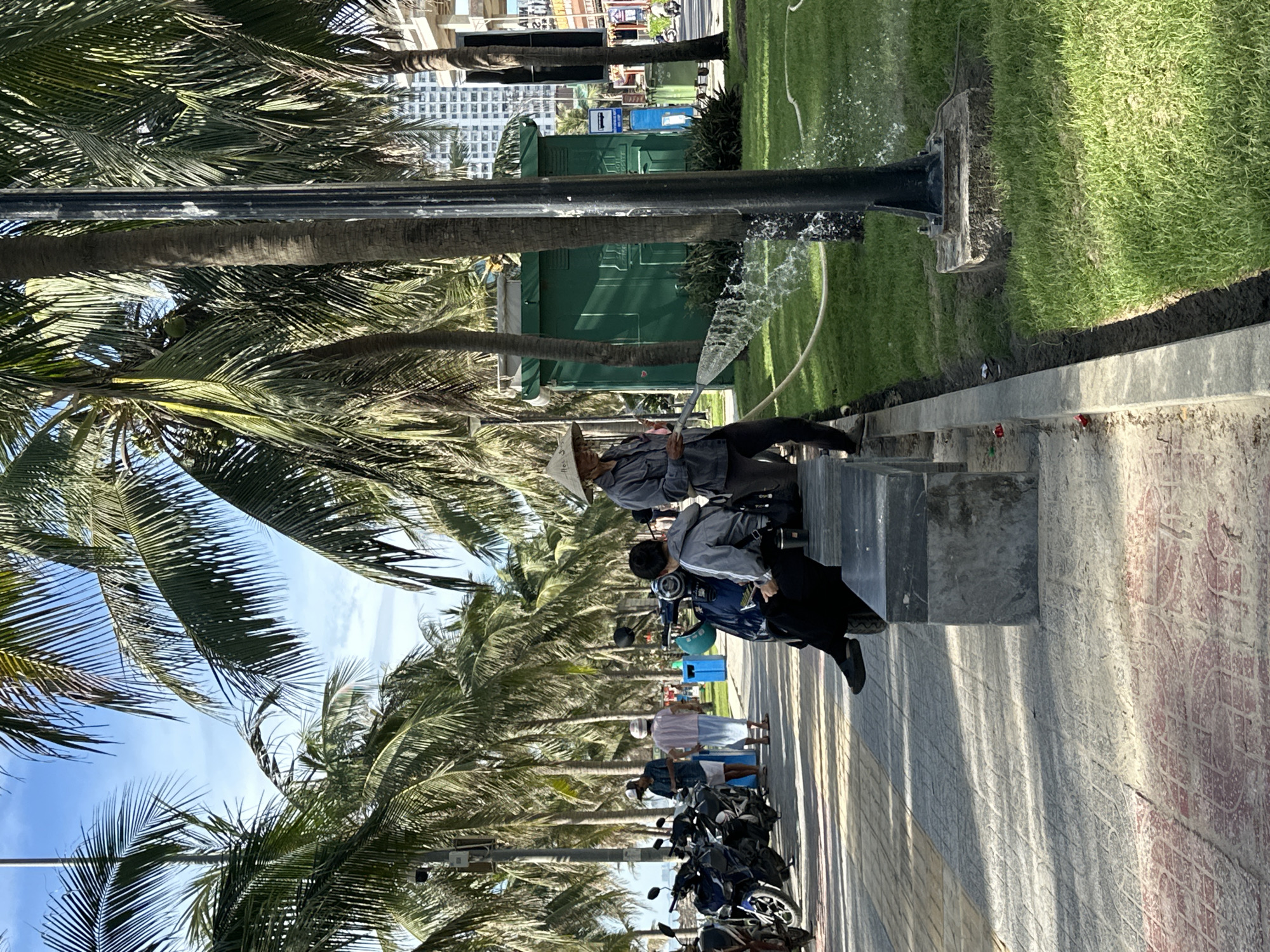 In Đà Nẵng, Vietnam, a man wearing a conical hat is watering grass with a hose, spraying water from a green portable toilet structure. He stands on a paved walkway next to a row of parked motorcycles, some of which are covered with tarps. To his right, a person sits on a concrete bench, facing away from the camera. In the background, palm trees line the street, and buildings are visible in the distance. The sky is a clear blue with some scattered clouds.
