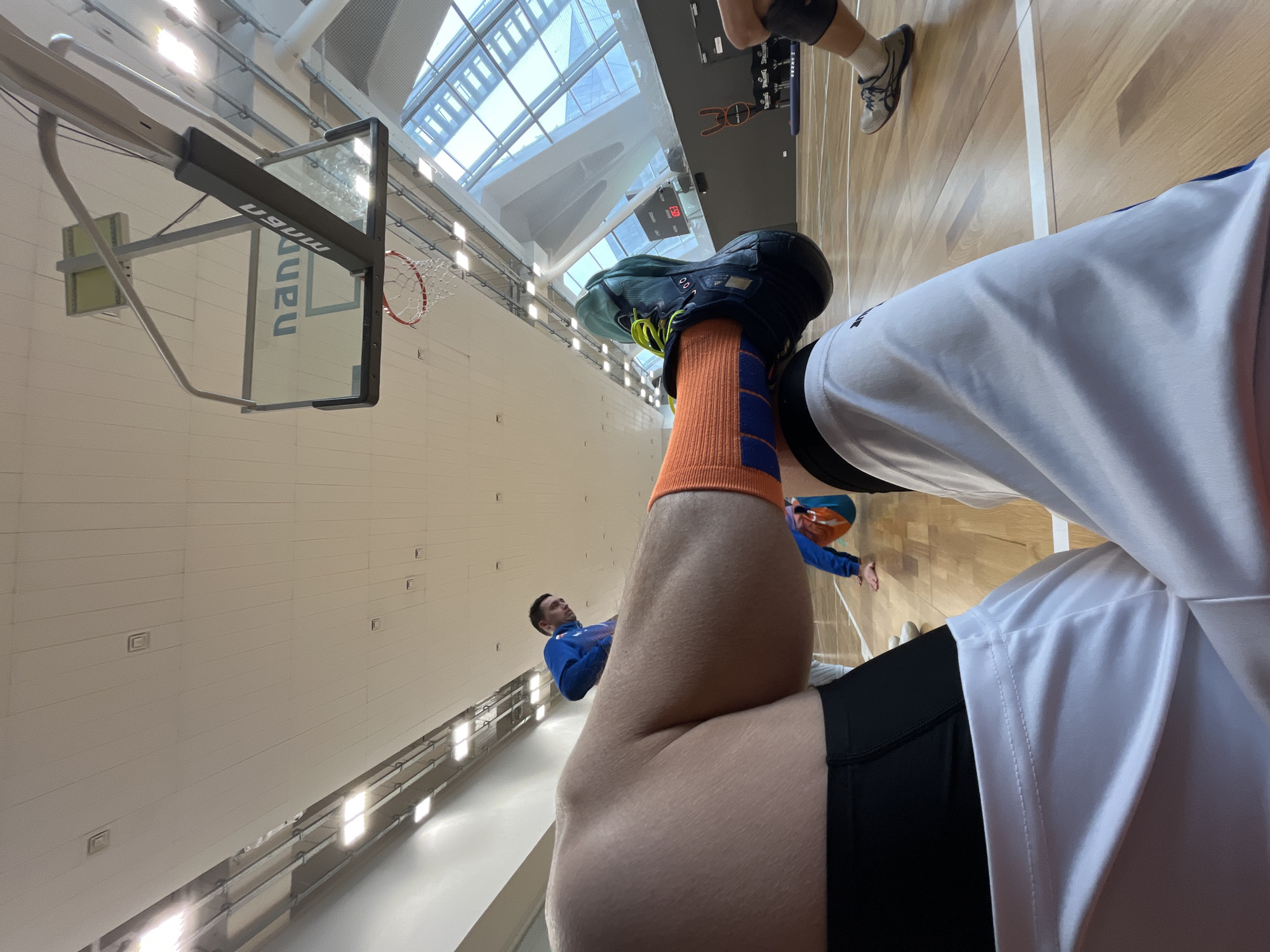 The image captures a scene inside a basketball gymnasium. The focus is on a person wearing athletic apparel, including a white jersey, black shorts, an orange sock with a blue stripe, and athletic shoes. The perspective is a low-angle shot, potentially taken by the person whose legs and torso are in the foreground.
In the background, another person is visible wearing a blue jacket. A basketball hoop is also visible. The gym has a high ceiling with numerous windows.
The image appears to be taken at the "MGUU Government University of Moscow" basketball court in the city of Moscow, Russia.