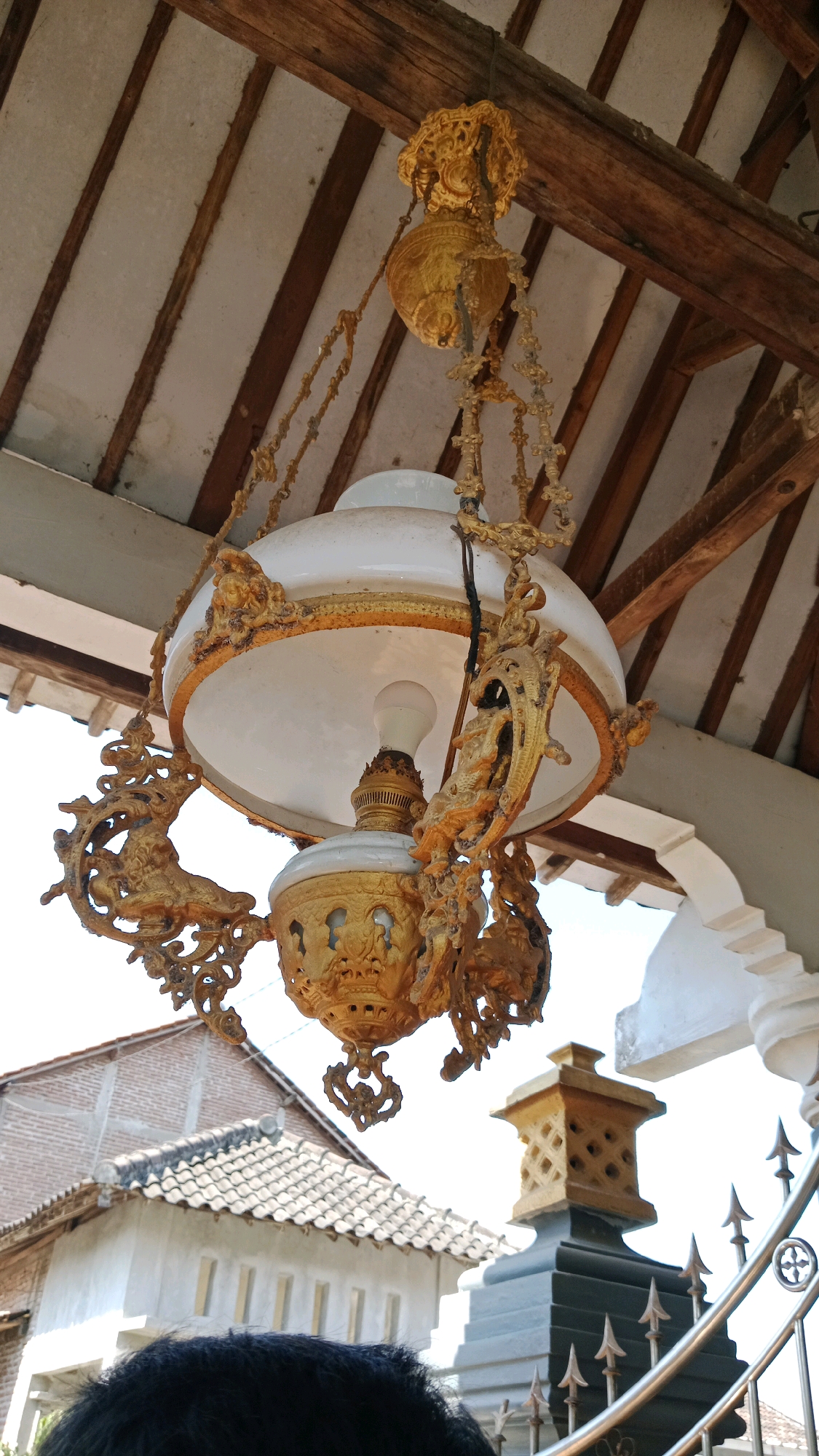 A detailed, ornate golden chandelier hangs from exposed wooden beams beneath a tiled roof structure, likely a veranda or outdoor pavilion. The chandelier features intricate scrollwork and decorative elements, with a large white lamp shade and a visible light bulb. Below the chandelier, part of a building with a tiled roof and brickwork is visible, along with a decorative stone post and a metal fence. A person's dark hair is visible in the foreground, suggesting they are standing directly beneath the chandelier, looking up. The scene appears to be outdoors, with bright daylight. The location is identified as Kinandang, Indonesia.