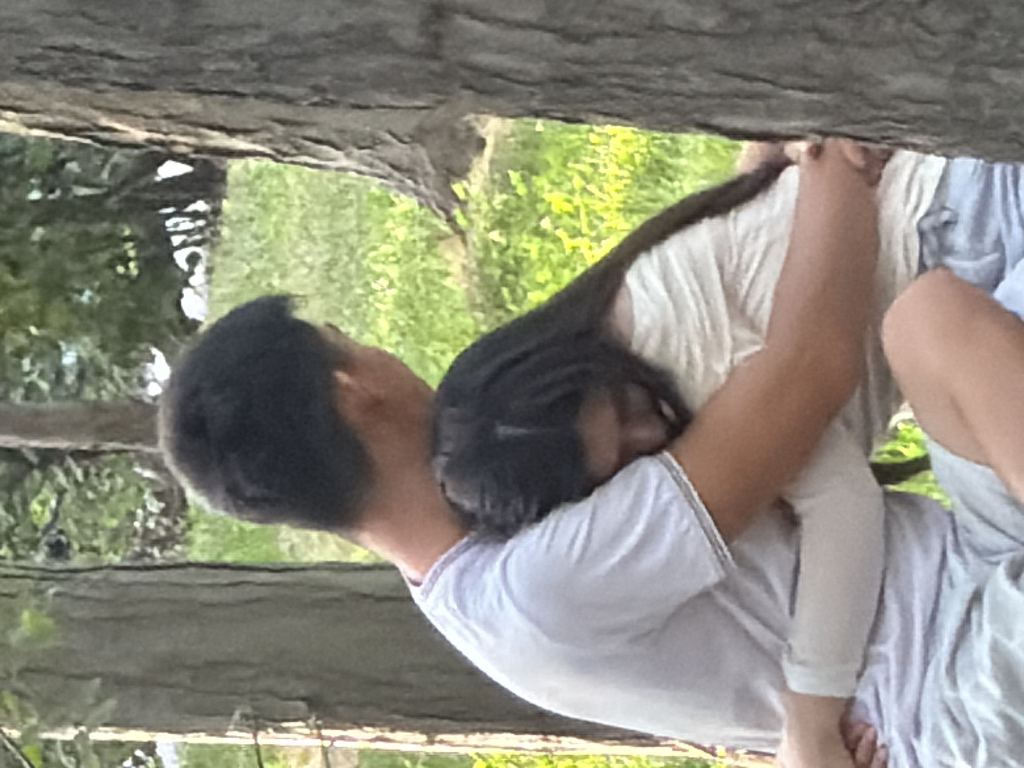 A young man and woman are embracing, seated outdoors, likely in a park or natural setting given the presence of trees and grass. The man, on the left, wears a light-colored t-shirt and is seen from the back, with his head tilted down as if looking at the woman. The woman, on the right, is also seen from the back, with her head resting on the man's shoulder. She has dark hair and wears a light-colored, off-the-shoulder top. Her arms are around the man. They appear to be in a moment of quiet affection. The background consists of green foliage and the textured bark of trees, suggesting an outdoor environment.