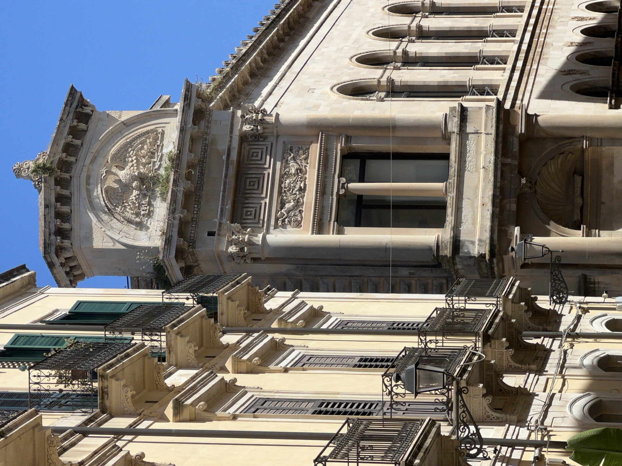 This is a close-up, low-angle exterior shot of an ornate building in Syracuse, Italy, likely in the historic district of Ortigia. The architecture displays classic Baroque elements with detailed stonework and wrought-iron balconies. On the left, a cream-colored building features multiple balconies with intricate black railings, green shutters on windows, and decorative corbels supporting the balconies. A street lamp with an ornate bracket is visible on one of the balconies. To the right, a more imposing stone building dominates the frame, showcasing a large, semi-circular relief sculpture of a bird or eagle surrounded by foliage, topped with decorative molding. This section of the building features classical columns and arched windows. The sky is a clear, bright blue, indicating a sunny day. No people are visible in the scene.