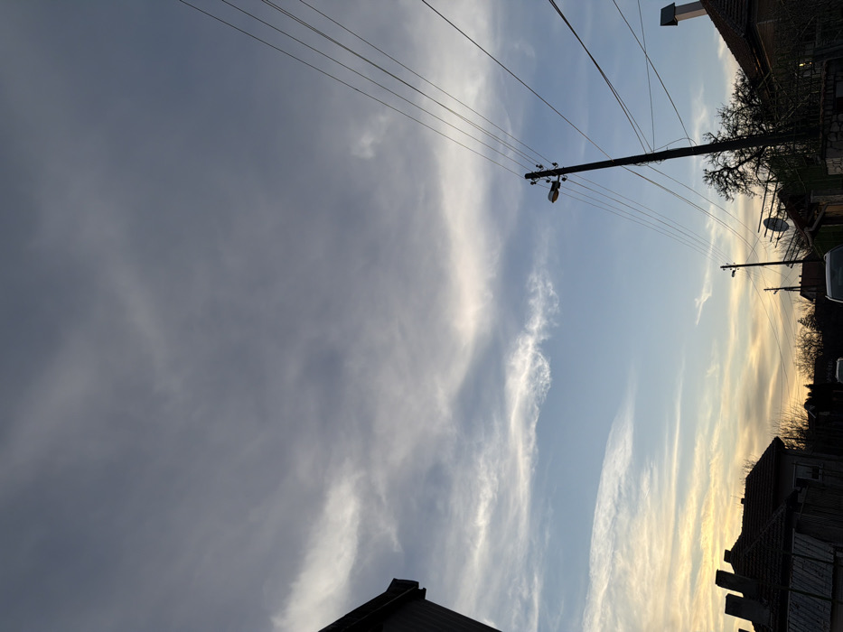 The image displays a residential street scene in Inovo, Bulgaria, observed at either dawn or dusk. The sky occupies the majority of the frame, transitioning from a dense layer of gray cloud cover in the upper half to a clearer, light blue sky with wispy white clouds and a distinct orange-yellow hue on the horizon to the left. Multiple utility poles, dark in silhouette, are present. A prominent pole in the mid-right features a single streetlight and supports numerous power lines that traverse diagonally across the frame. Additional poles and power lines extend into the background. Residential structures with dark-tiled roofs and chimneys are visible on both sides of the street, largely silhouetted against the horizon. Deciduous trees, appearing bare, are interspersed among the buildings and utility infrastructure. Several dark vehicle shapes are faintly discernible on the unlit street in the distance. No individuals or active interactions are present in the scene.