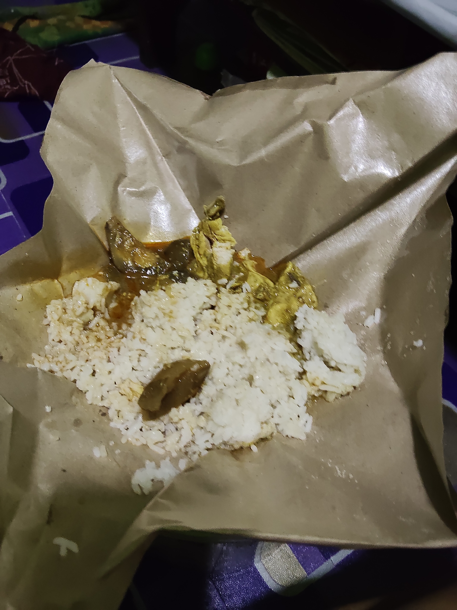 The image shows a close-up overhead view of a meal served on brown paper. The meal consists of white rice with some sauce and pieces of meat or poultry. A dark brown, somewhat oval-shaped object is visible in the center of the rice. The food appears to be partially consumed. The brown paper is crumpled and forms a makeshift serving base. The background is out of focus, but suggests a table with a patterned surface, possibly a purple and white checkered design. There are no people or specific landmarks visible that would indicate a precise location within Lori, Indonesia.