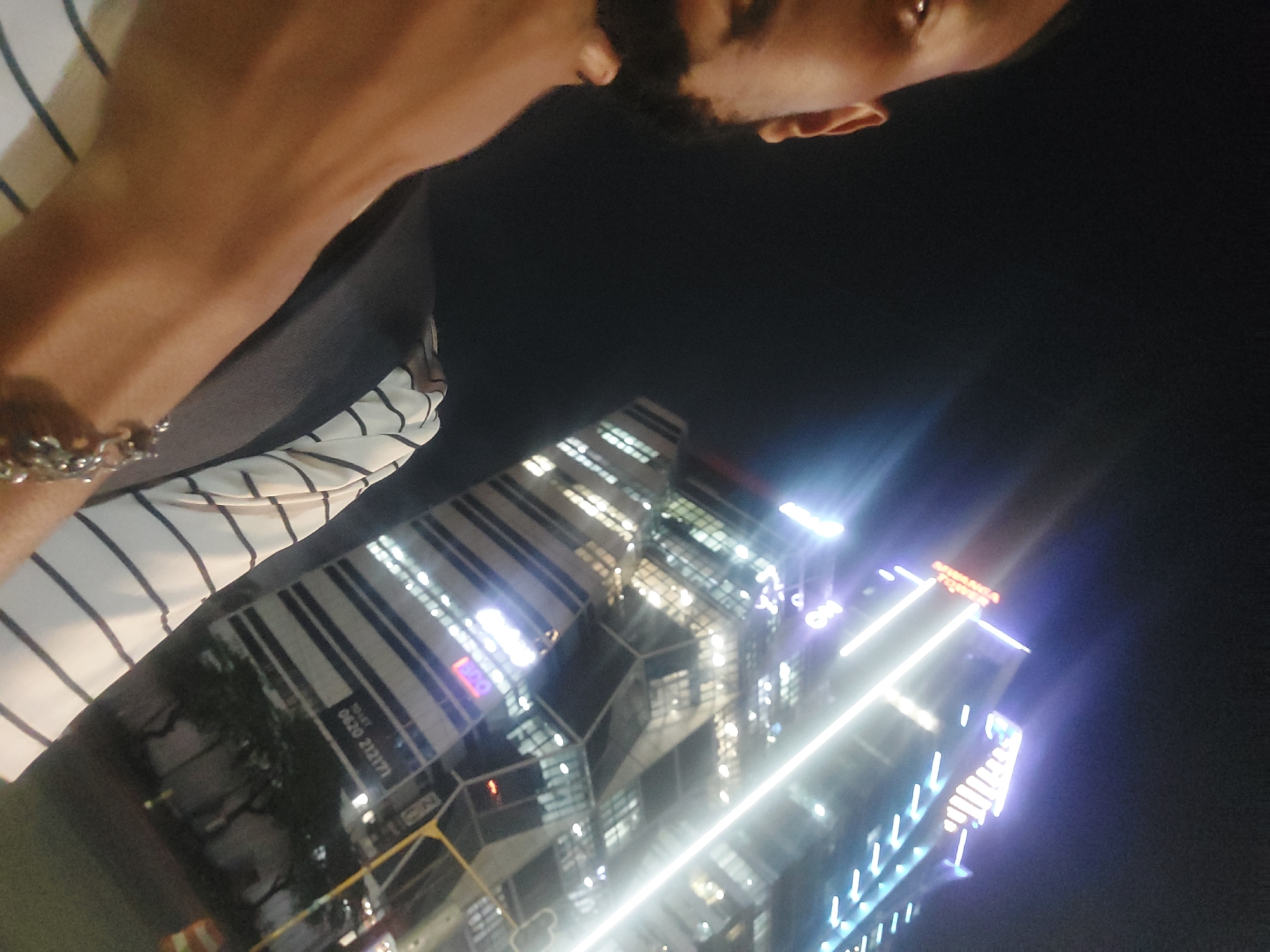 A low-angle shot features a person from the chest up, wearing a striped shirt, with a cityscape at night in the background. The person has dark hair and facial hair. The buildings are illuminated with lights, including some with prominent vertical light bars. Text can be seen on one building, along with a visible phone number. The environment suggests a modern urban setting. While I can identify the location as Dar es-Salaam, Tanzania, from associated information, it is not possible to conclusively identify it from visual features in this image alone.