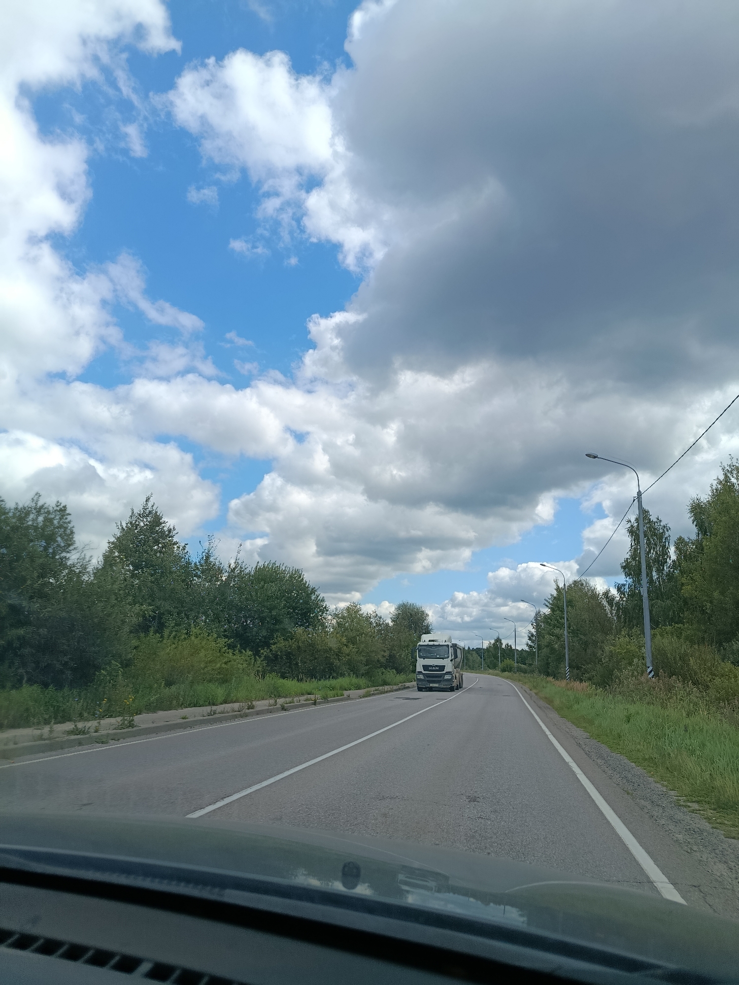 The image captures a view from a vehicle traveling on a paved road in Savinskaya, Russia. The road has a white dashed line marking the center and a solid white line on the right edge. A white truck with the logo "MAN" is visible in the distance, driving on the road. The environment is characterized by lush green trees and vegetation lining the sides of the road. The sky above is a mix of blue sky and dark, dramatic clouds. Several streetlights are positioned along the road. The foreground shows the interior of the vehicle from which the photograph was taken.