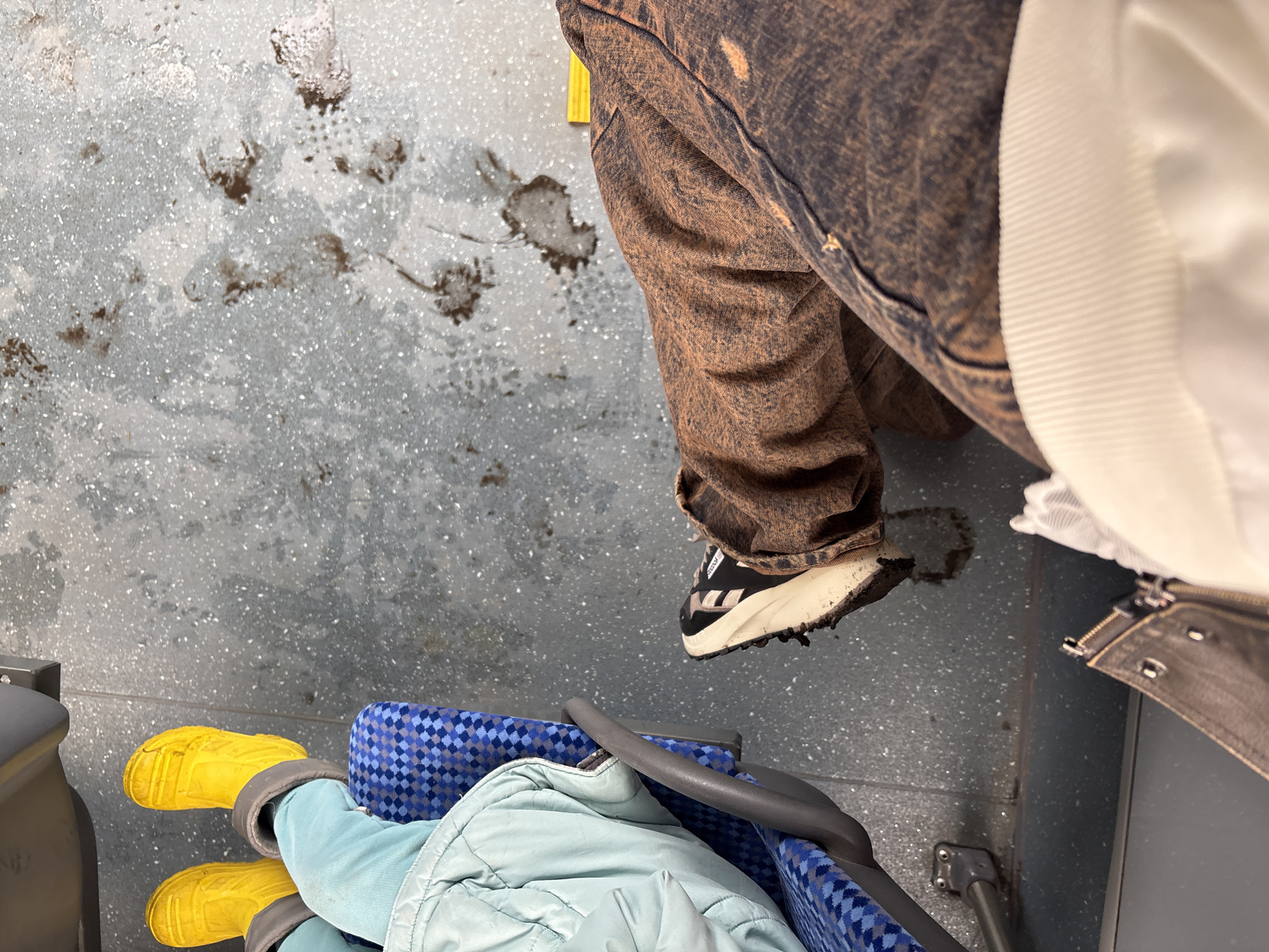 The photograph, taken from a high angle, depicts the floor of what appears to be a public transport vehicle, possibly a train, due to the patterned blue seat and grey floor. The floor is stained and appears dirty. The lower part of a person wearing brown denim pants and black and white sneakers is visible in the upper right quadrant. Another person, likely a child, is sitting in the seat with yellow boots, a light blue jacket, and a blue patterned seat. The image provides no direct evidence to place the photograph in Krasnoyarsk, Russia.