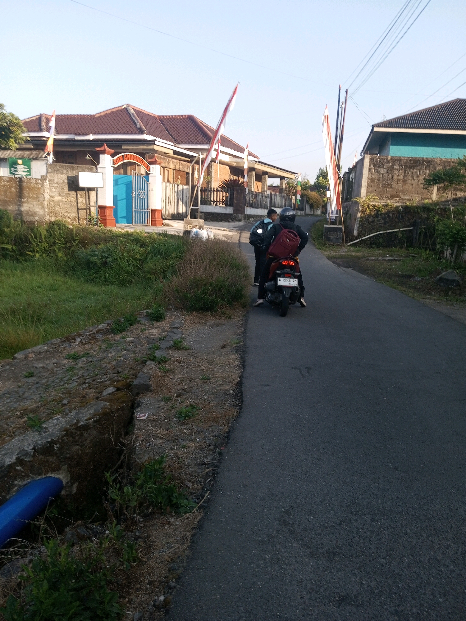 In Andongsili, Indonesia, two individuals are captured on a paved road. The person in the foreground is riding a red and black motorcycle, wearing a black helmet and a maroon backpack. They are traveling away from the camera. To their left, another person, likely a passenger or companion, stands on the road's edge, also wearing a backpack. The road is flanked by grassy areas and a low concrete wall on the left, with a blue pipe visible emerging from the wall. On the right, a building with a turquoise roof and stone walls is visible, along with another structure further back that appears to be a residential house with a brown tiled roof. Indonesian flags are prominently displayed, attached to poles along the roadside in front of the buildings. The sky is clear and blue.