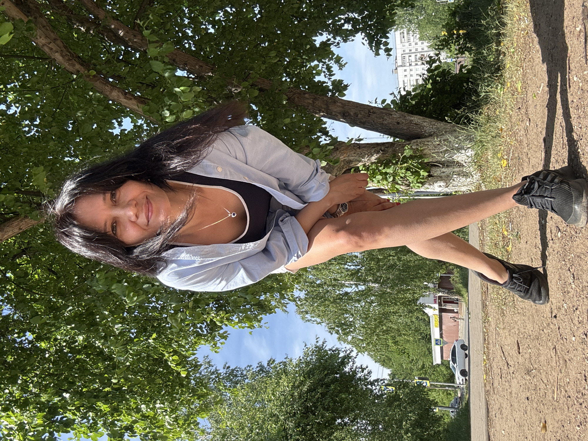 A full-length, outdoor photograph captures a woman under the shade of trees in what appears to be a park or green space within a city. She is positioned at an angle, facing the camera with a slight tilt. She wears a black top under a light blue, button-down shirt, and dark athletic shoes. Her dark hair is loose, and she wears a necklace.
The setting features lush green trees overhead, creating a canopy, with a glimpse of a blue sky in between the leaves. In the background, there is a building. A path runs alongside where the woman is sitting, and a road with parked cars and a building are visible further in the background. The ground appears to be a mix of dirt and vegetation.