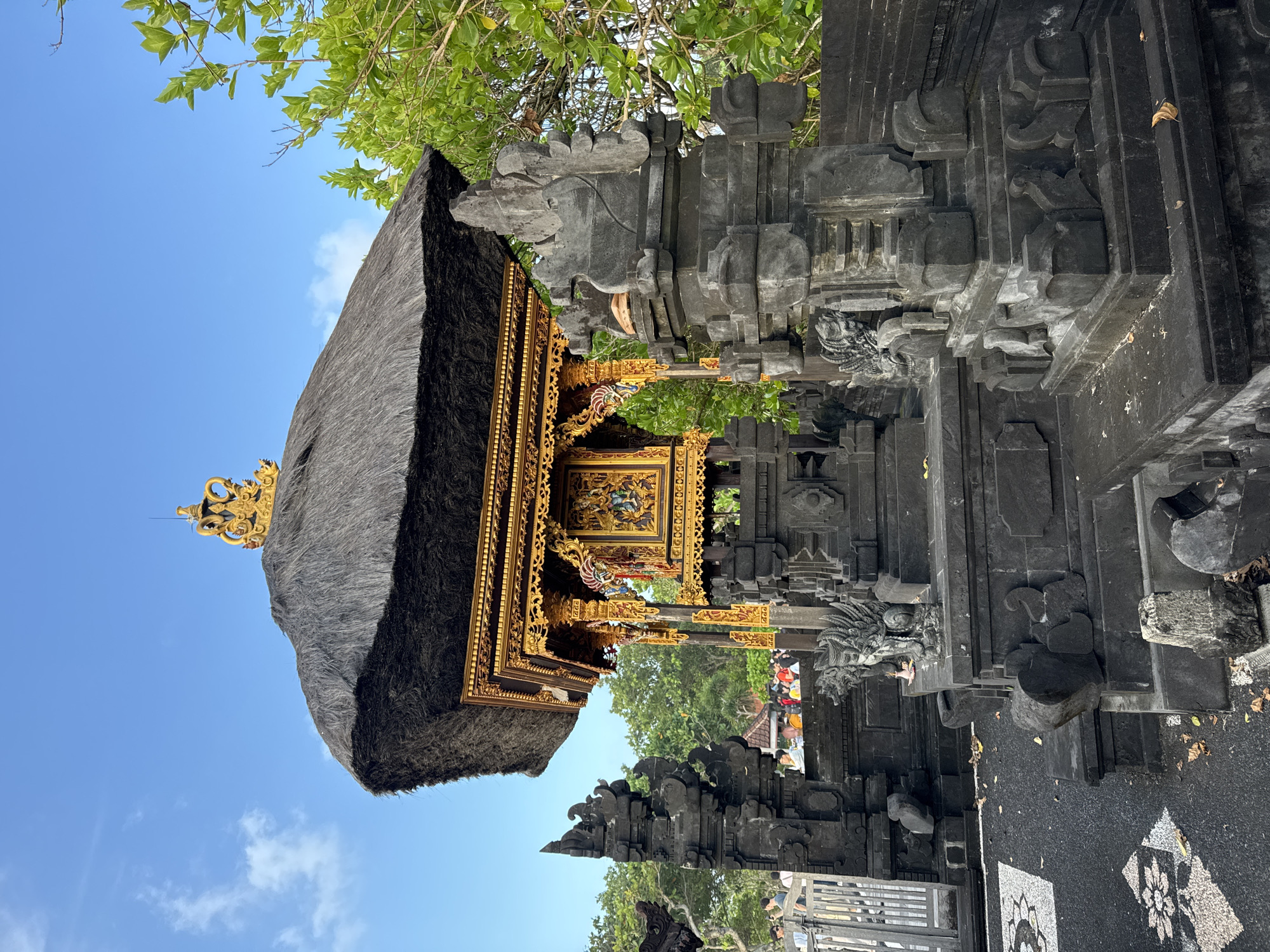 The image shows a Balinese Hindu temple structure in Kediri, Indonesia. The building features a traditional thatched roof with a decorative gold finial. Intricate gold detailing adorns the wooden structure beneath the roof. The building is framed by carved dark grey stone structures, possibly part of a gateway or compound wall. The sky is a light blue with some clouds and green foliage is visible. People with colored heads can be seen through the gate. The ground is covered in black asphalt, along with some decorative tiles, and fallen leaves.