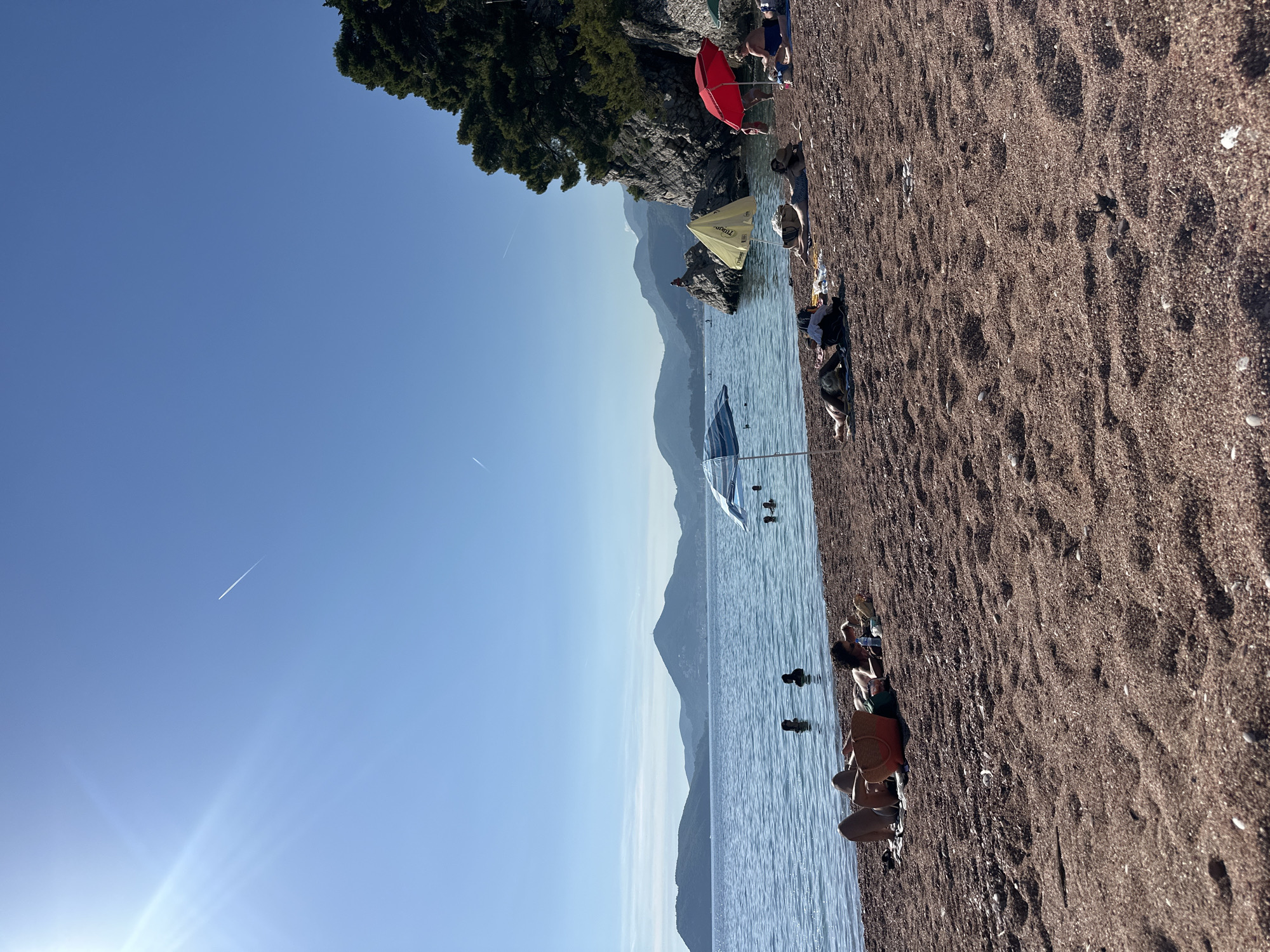 A beach scene in Sveti Stefan, Montenegro, shows several people sunbathing and swimming under a clear blue sky. The beach is covered in reddish-brown pebbles. In the water, several individuals are swimming, and further out, mountains form a hazy backdrop. There are at least three beach umbrellas providing shade for people on the shore. One person is seated under a red umbrella, while another is lying down near a yellow umbrella. A striped umbrella stands further to the left. The sun is shining brightly, casting a lens flare in the upper left corner of the image. A faint airplane contrail is visible in the sky. Dense green trees and rocky outcrops are present on the right side of the image, extending towards the water.