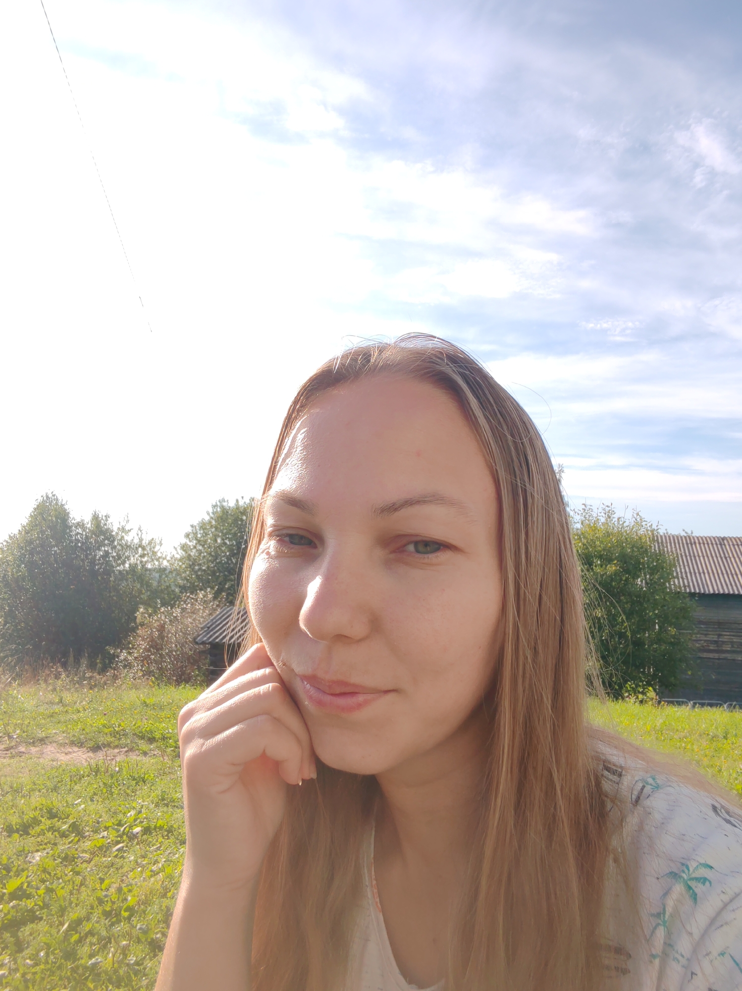 A young woman with long blonde hair is taking a selfie outdoors. She is resting her chin on her hand and smiling slightly. The background shows a grassy field, trees, a wooden building, and a blue sky with some clouds. The overall lighting suggests it is daytime. The location is in or near the city of Chukhlem, Russia.