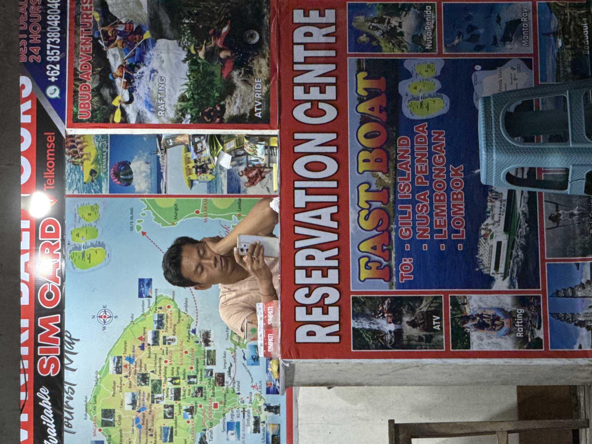 The image shows a person holding a phone in front of a promotional sign for tourist activities, possibly in North Kuta, Indonesia. The sign advertises "Fast Boat" trips to Gili Island, Nusa Penida, Lembongan, and Lombok, along with other attractions like rafting and ATV rides. It also promotes "Ubud Adventures" and advertises SIM cards for tourists. The person holding the phone appears to be looking at the screen and is positioned in front of a tourist map of Bali. There is a blue plastic stool nearby and other objects suggesting an indoor environment.