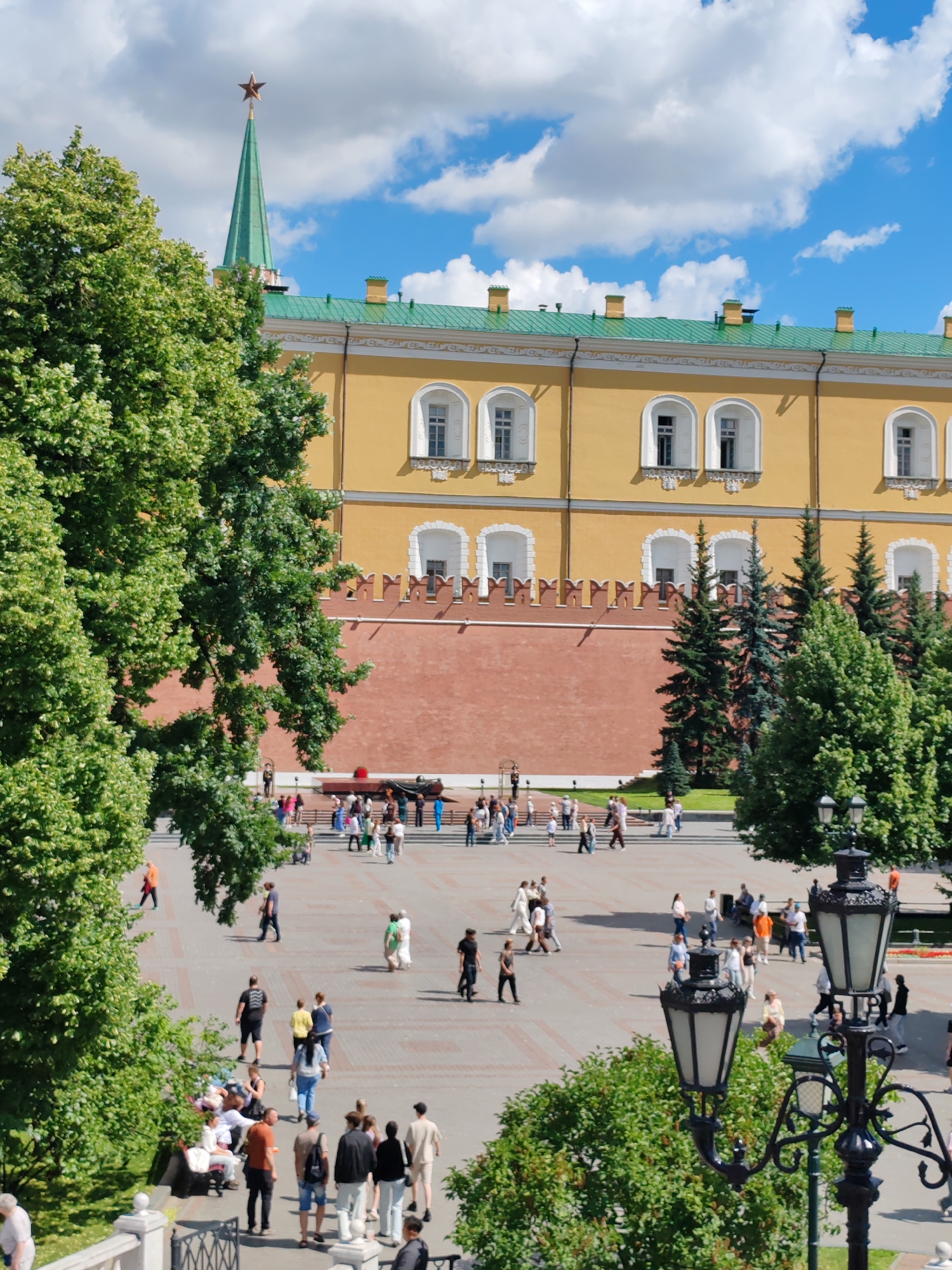 An elevated, eye-level shot captures a bustling square in front of a grand, yellow building. The building, with its green roof and red brick lower section, is adorned with arched windows and crenellations. A tall green spire topped with a red star pierces the blue sky, scattered with white clouds. The square itself is paved with light-colored stones and is populated by a diverse crowd of people walking, standing, and sitting. Some individuals are dressed in casual summer attire. Lush green trees frame the scene on the left and right. Two ornate black lampposts stand in the foreground, adding to the ambiance of the urban setting. The overall impression is one of a lively public space on a bright, sunny day.