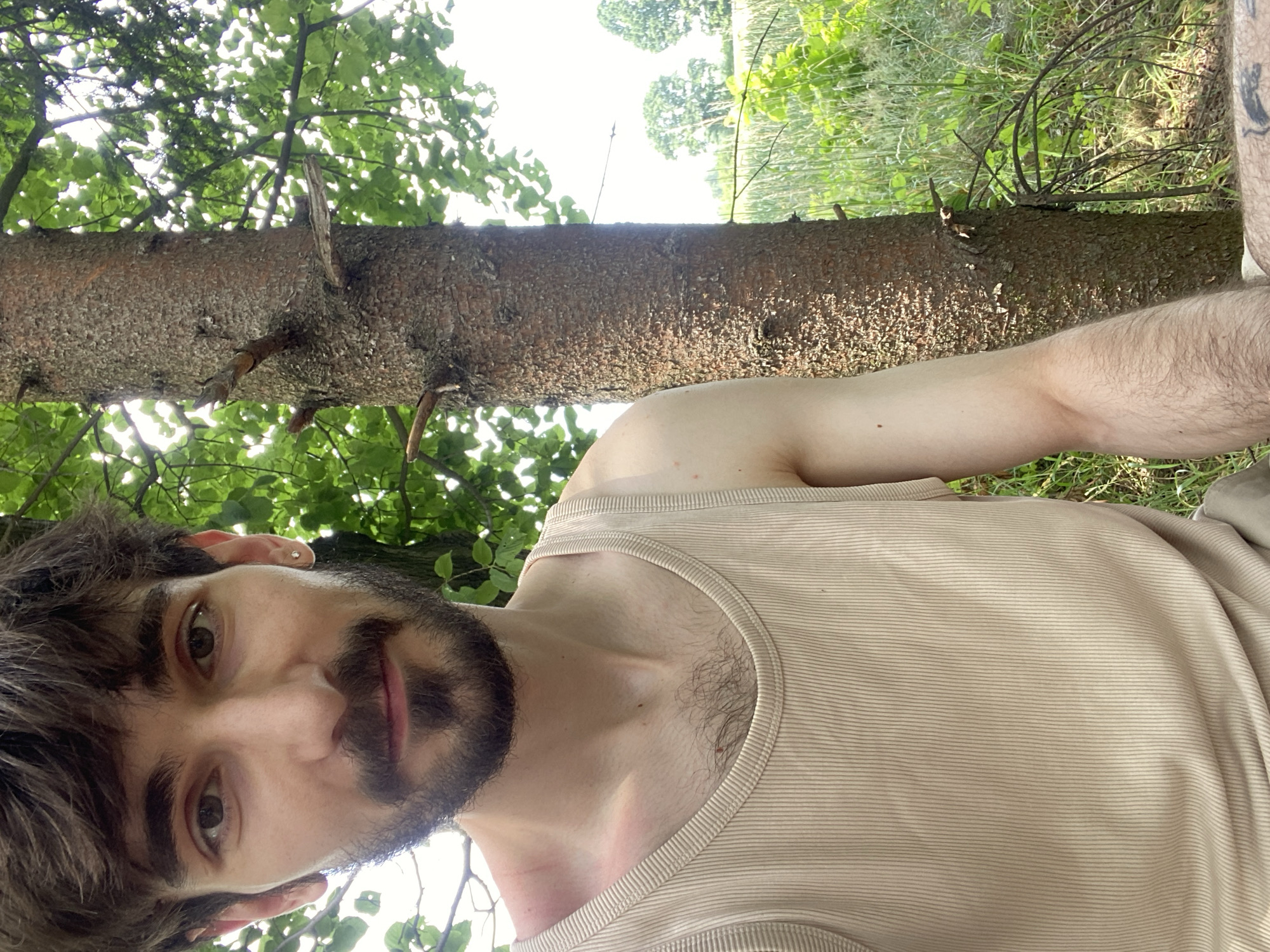 The image is a selfie taken outdoors, likely in Kaniów, Poland, given the context. A young man with brown hair and a beard is positioned in the foreground, looking directly at the camera. He is wearing a beige ribbed tank top. To his right, a thick tree trunk is visible, with some branches extending out. The background consists of lush green foliage and grass, with a hint of a field and distant trees. The lighting suggests it is daytime, with sunlight filtering through the leaves. The man's left arm is visible, and a tattoo can be seen on his left thigh. The overall scene depicts a casual outdoor setting.