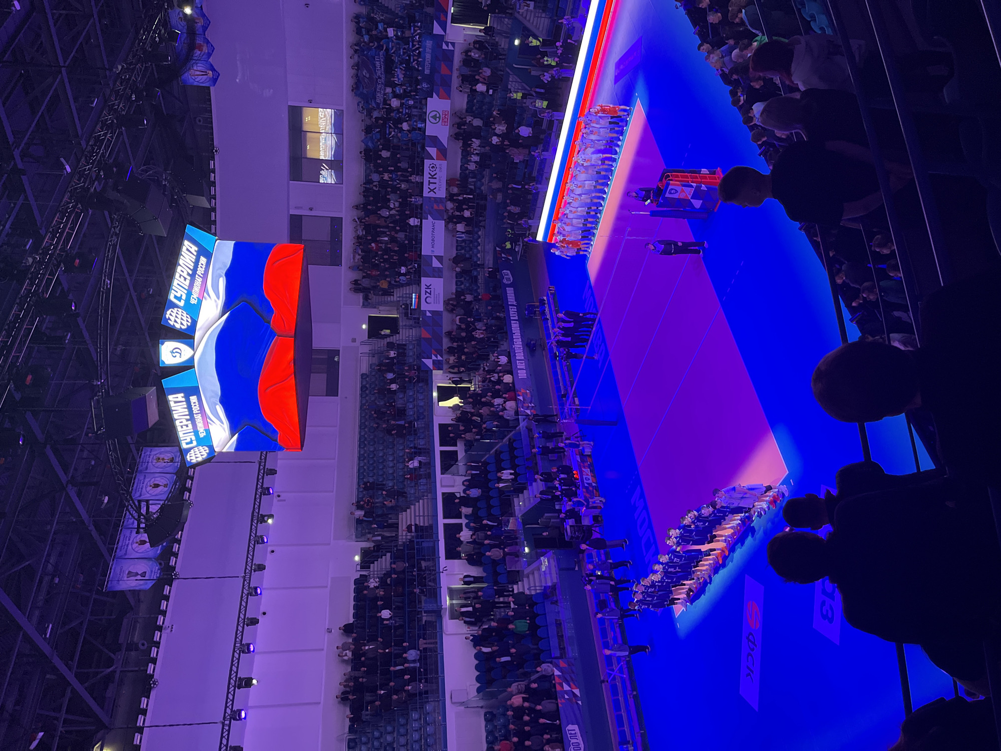 The image shows an indoor volleyball game in progress at the Dynamo Volleyball Arena in Moscow, Russia. The stands are filled with spectators. A large video screen displays the Russian flag and the word "Superliga" in Russian, which is the name of the Russian volleyball league. The volleyball court is brightly lit. The volleyball teams are lined up on either side of the court, facing each other.