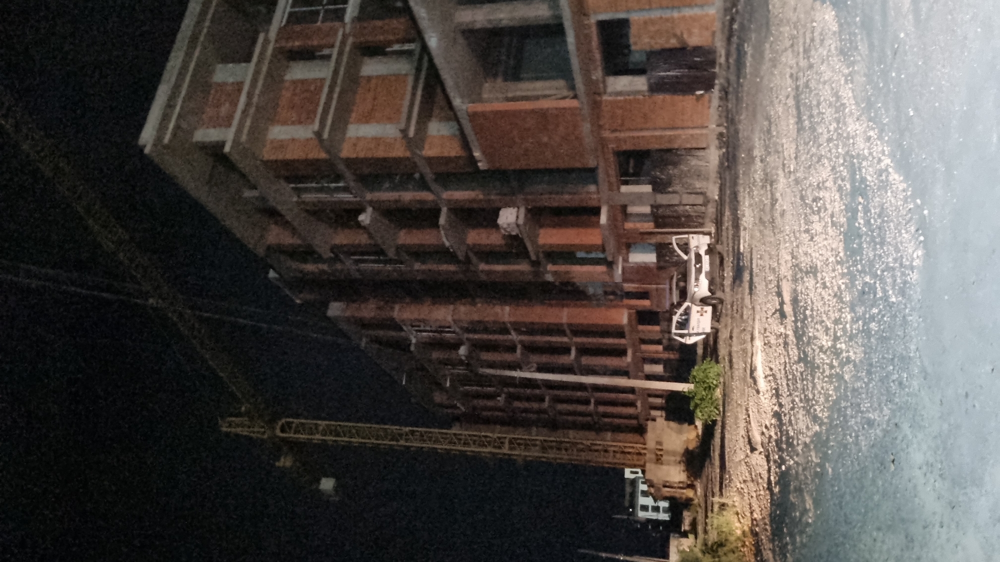 An external, low-angle, nighttime shot depicts an unfinished brick building under construction in Mukachevo, Ukraine. A large yellow construction crane is positioned to the left of the building, its boom extending across the frame. A white utility vehicle, possibly a police car, is parked in front of the building with its doors open. The ground in the foreground is unpaved and covered in gravel and debris. The sky is dark, indicating it is nighttime. The image appears to be taken from a street or construction site.