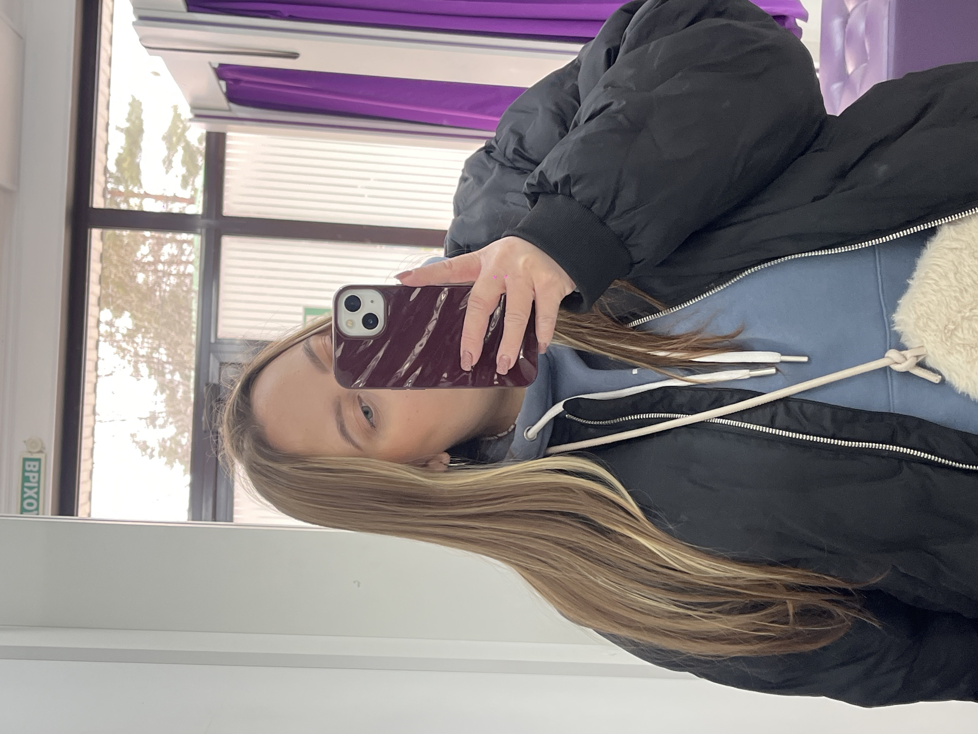 A person is holding a cell phone in front of their face, taking a picture in what appears to be a fitting room or a shop with a window. The window shows a view of an exterior location that can be recognized as Novopokrovskaya, Russia, from the sign visible outside. The person is wearing a black jacket and a blue hoodie. The fitting room has purple accents in the curtains.