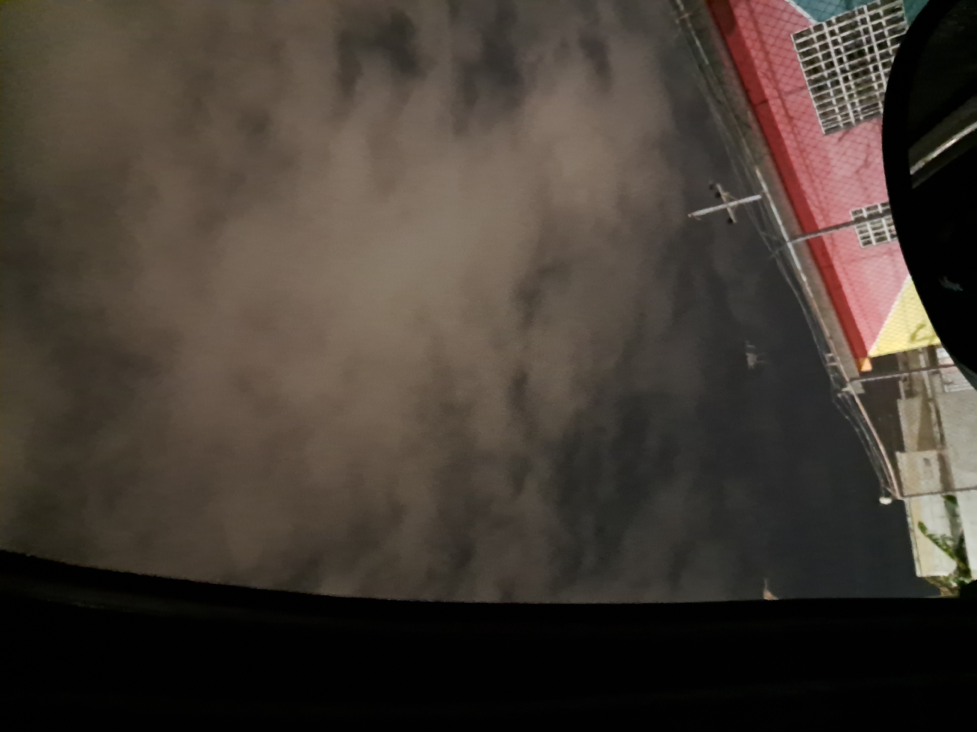 A nighttime exterior shot from inside a vehicle in Cumana, Venezuela, shows a multi-colored building with a red and yellow facade, featuring a white grid-like window. A utility pole with wires is visible in front of the building. The sky is filled with dense, dark clouds, illuminated by an unseen light source.