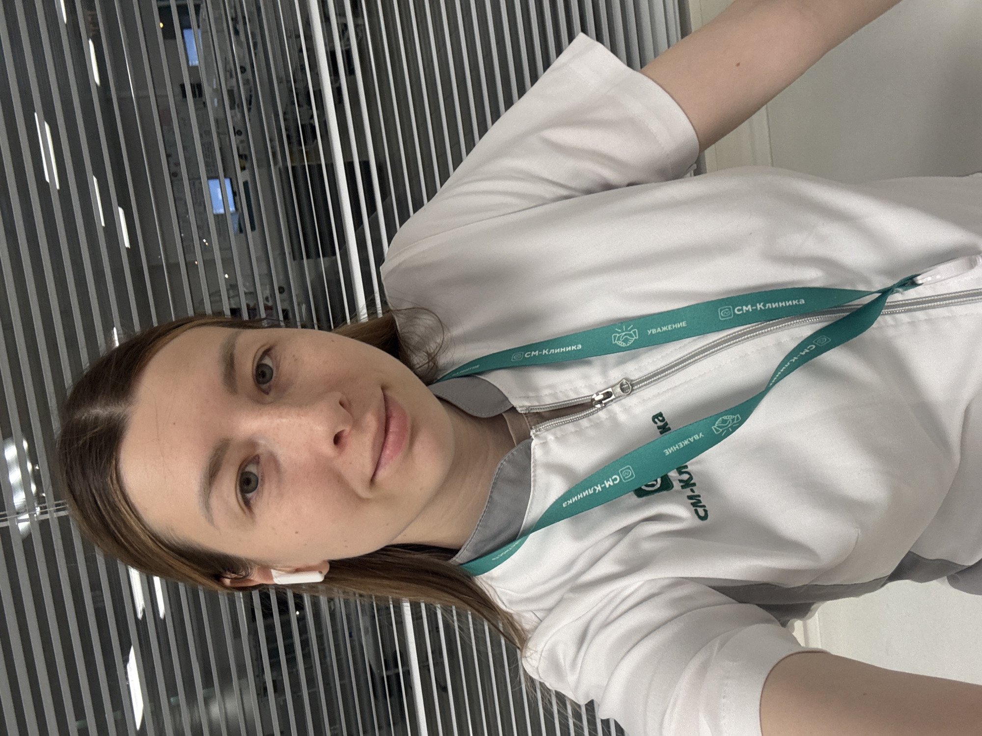 A selfie of a woman wearing a white medical jacket with a green lanyard that reads "СМ-Клиника" (SM-Clinic), possibly taken indoors near a window with blinds.