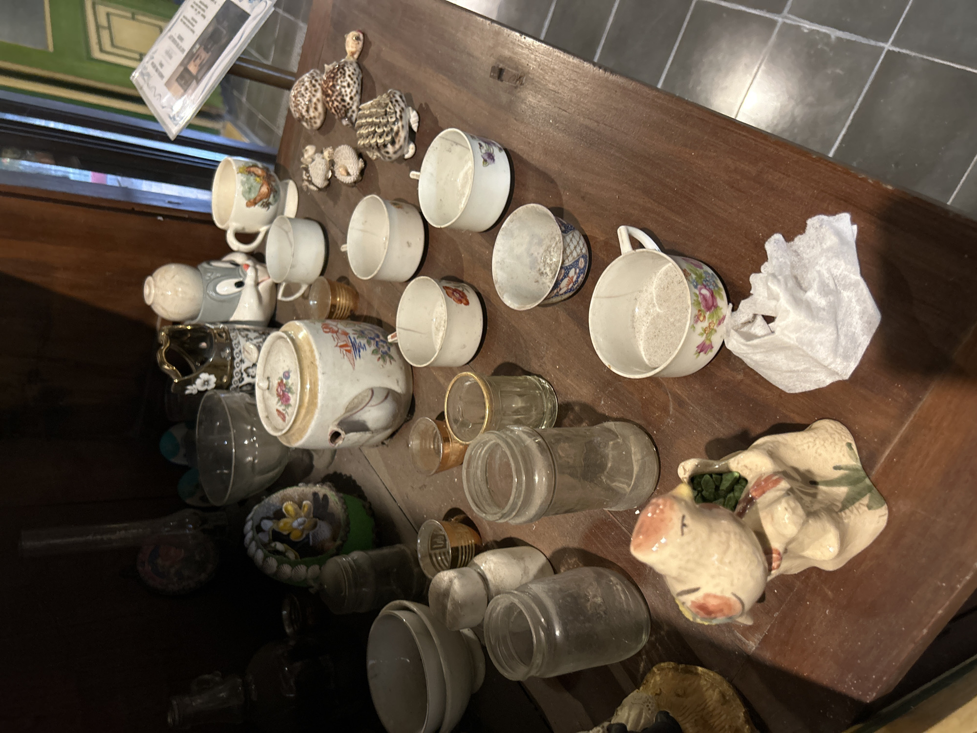 The image shows a collection of ceramic decorative items and tableware displayed on a dark wooden table. In the foreground, there is a ceramic figurine of a bird with green elements in its beak, and to its right, a crumpled white tissue. Further back on the table are several white ceramic cups and bowls, some with floral patterns. There are also glass jars and small decorative shells resembling turtles. On the left side of the table, there is a larger ceramic pot or teapot with a floral design and a handle. In the background, various other decorative objects are visible, including what appears to be a glass vase and a glass bowl. The overall setting suggests an indoor display, possibly in a shop or home, with a tiled floor visible in the lower right corner. This image is of a display of decorative ceramic items in a shop located in Banguntapan, Indonesia.