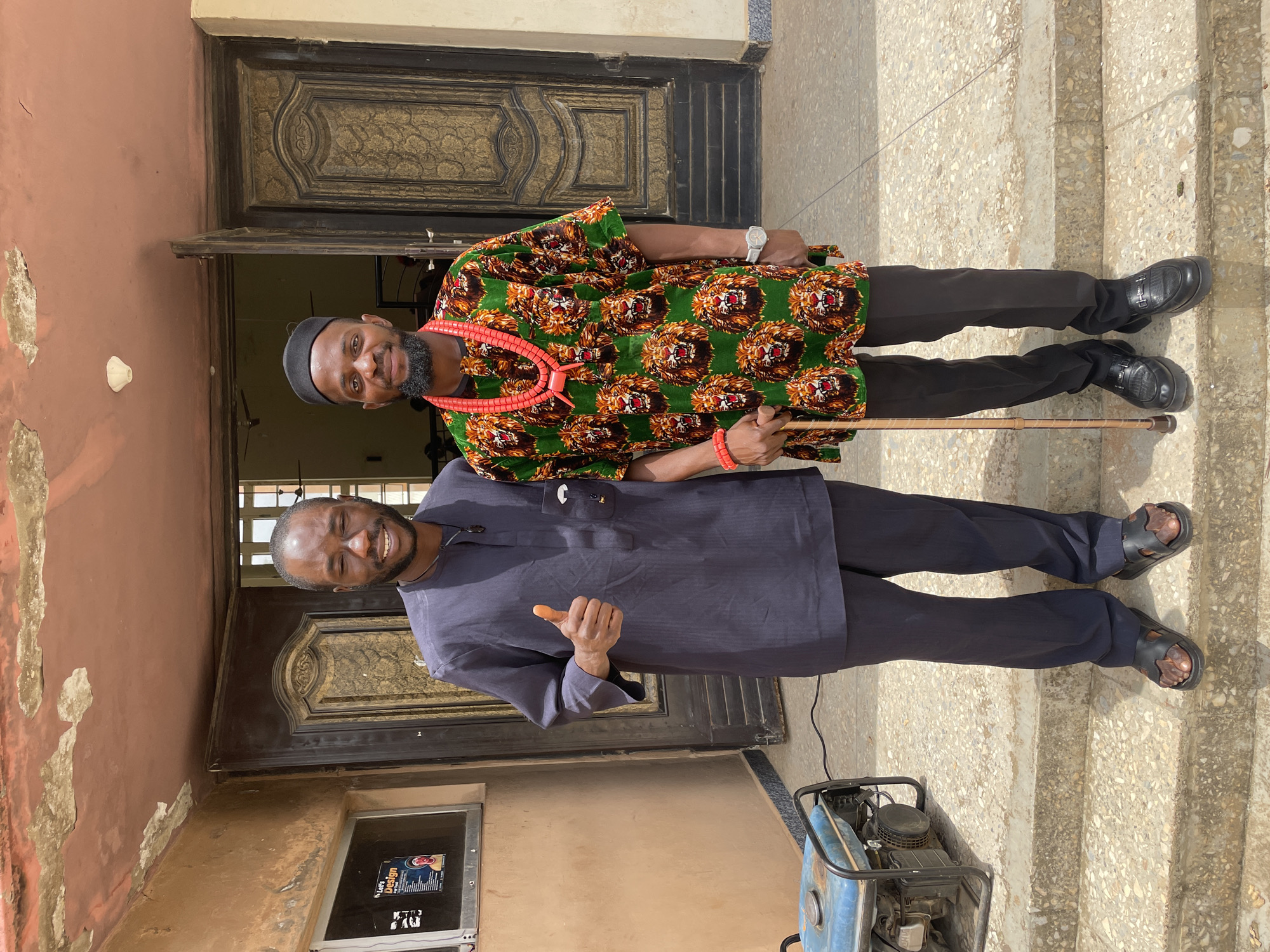 Two men are standing on steps outside a building in Jos, Nigeria. The man on the left is wearing a dark blue traditional outfit and giving a thumbs up. The man on the right is wearing a vibrant green shirt with a lion print, a red beaded necklace, a black cap, and holding a cane. A blue generator is visible to the left of the steps. The building appears to have a textured, peach-colored facade and a large, ornate dark brown wooden door. The sky is visible and appears to be overcast.
