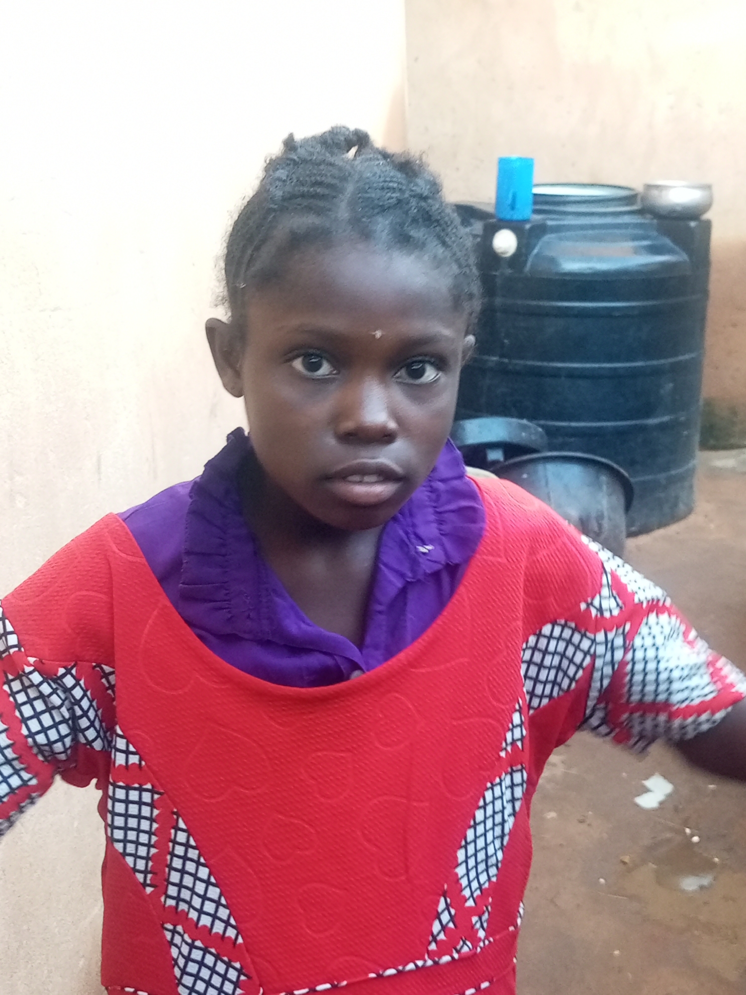 A young African girl with dark skin and braided hair is pictured in a medium close-up shot, looking directly at the camera. She is wearing a vibrant red dress with a purple collar. The dress features a pattern of black and white squares and scalloped edges. In the background, a large black water tank is visible, along with a smaller dark bucket. To the left, a light-colored wall provides a backdrop. The scene appears to be outdoors, possibly in a residential area or compound in Tempure, Nigeria, given the presence of water storage and common building materials. The lighting suggests it is daytime.
