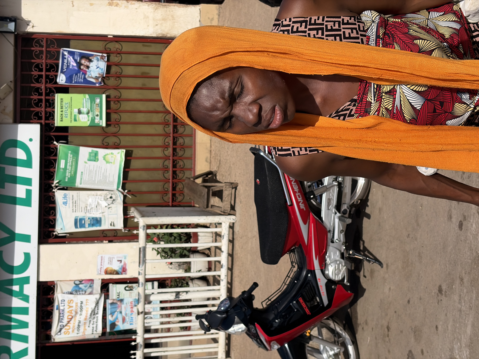 The image captures a woman in Gura Top, Nigeria, wearing an orange headwrap and a patterned top, positioned near a red and black Jin Cheng motorcycle and a pharmacy storefront labeled "PHARMACY LTD." The pharmacy has posters and advertisements on its facade, including signs for "REFUCIL", "VENILLE", "4Life", and information regarding "ED PHARM LTD" business hours for Sundays (1:00 PM - 9:00 PM), a phone number, and the “Junction Branch.” The environment consists of a paved street with a simple stool and a flower-filled white railing next to the building.