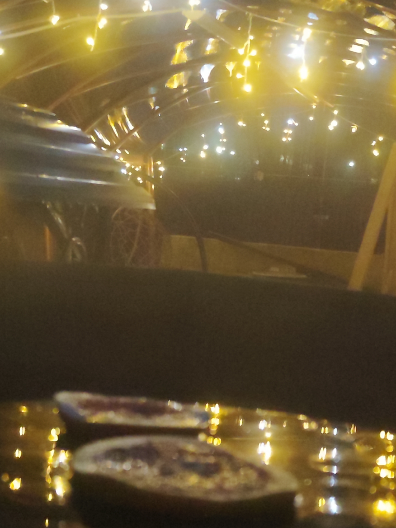 The image shows a blurry, low-lit scene with two small, round objects in the foreground, possibly food items with a reddish center and a light-colored crust. These appear to be sitting on a reflective surface illuminated by warm, yellow light sources. In the background, vague shapes suggest an interior space with architectural elements such as arches or beams decorated with strings of lights. A section of a vehicle is visible on the left of the center. Given the specified location of Chaikovsky, Russia, this could potentially be a cafe or restaurant with outdoor seating, where the food items are being served and the reflective surface is a table. The overall impression is one of a dimly lit, perhaps festive, atmosphere.