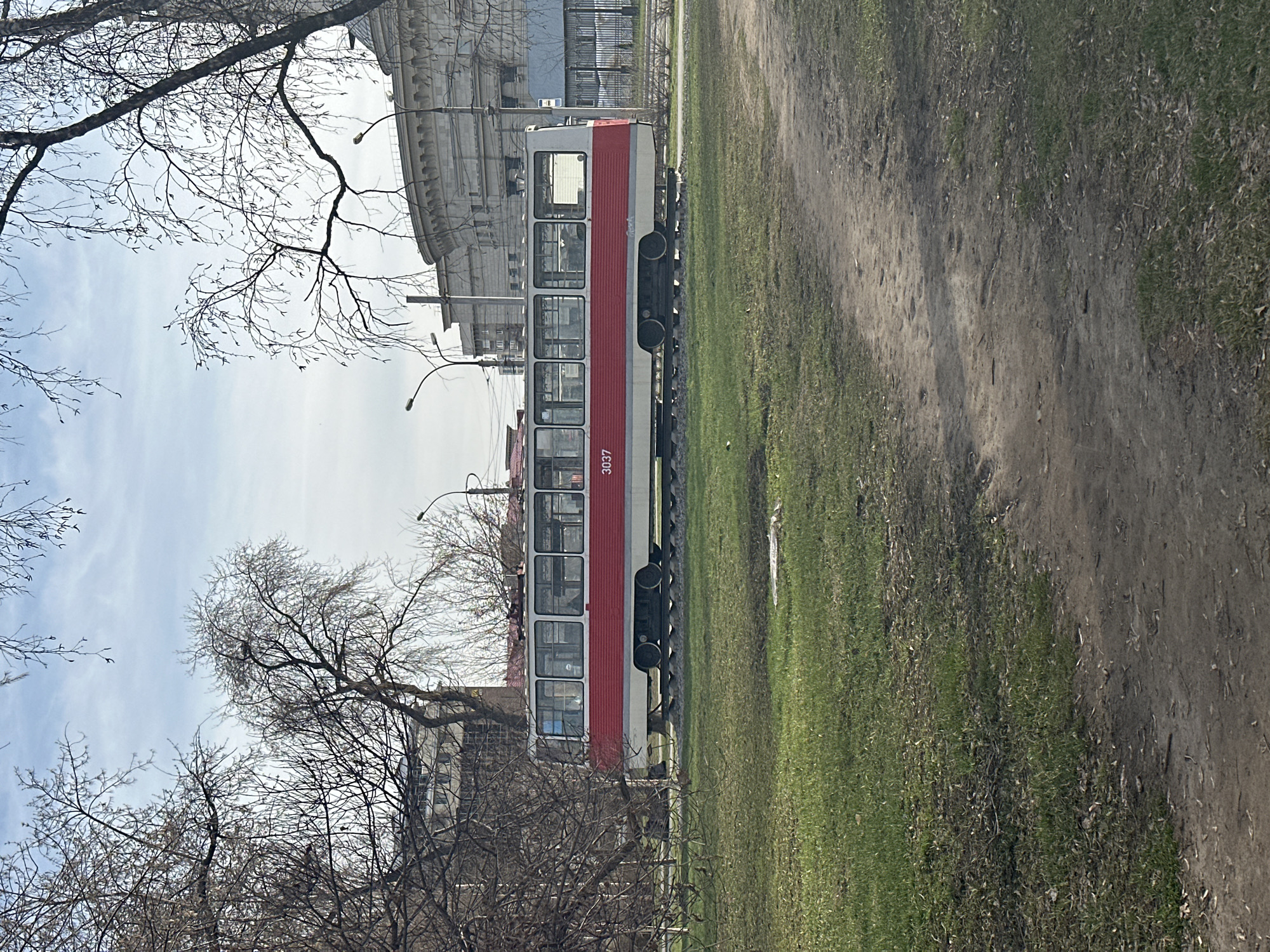 A red and white tram with the number 3037 is parked on a railway track. The tram is positioned next to a patch of grass and a dirt path. In the background, there is a building with architectural detailing and bare trees. The sky is partially cloudy. The image was taken in Novosibirsk, Russia.